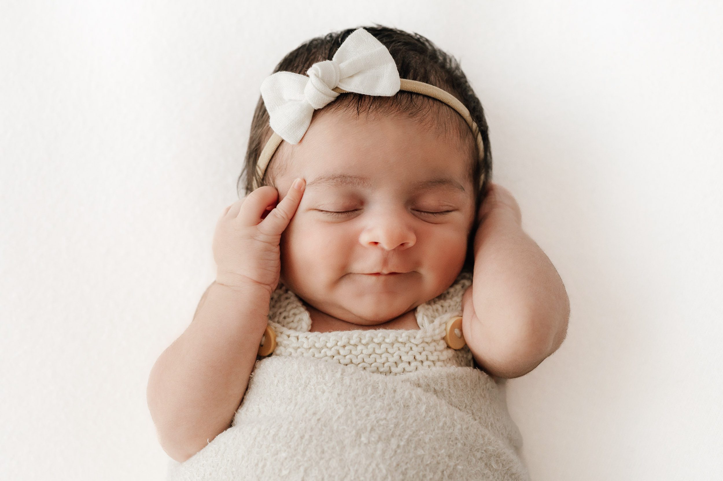 a newborn baby girl wrapped in a fuzzy swaddle blanket sleeping on a white backdrop as she holds her hands up to her cheeks with a hint of a smile on her face during a natural light newborn photo session