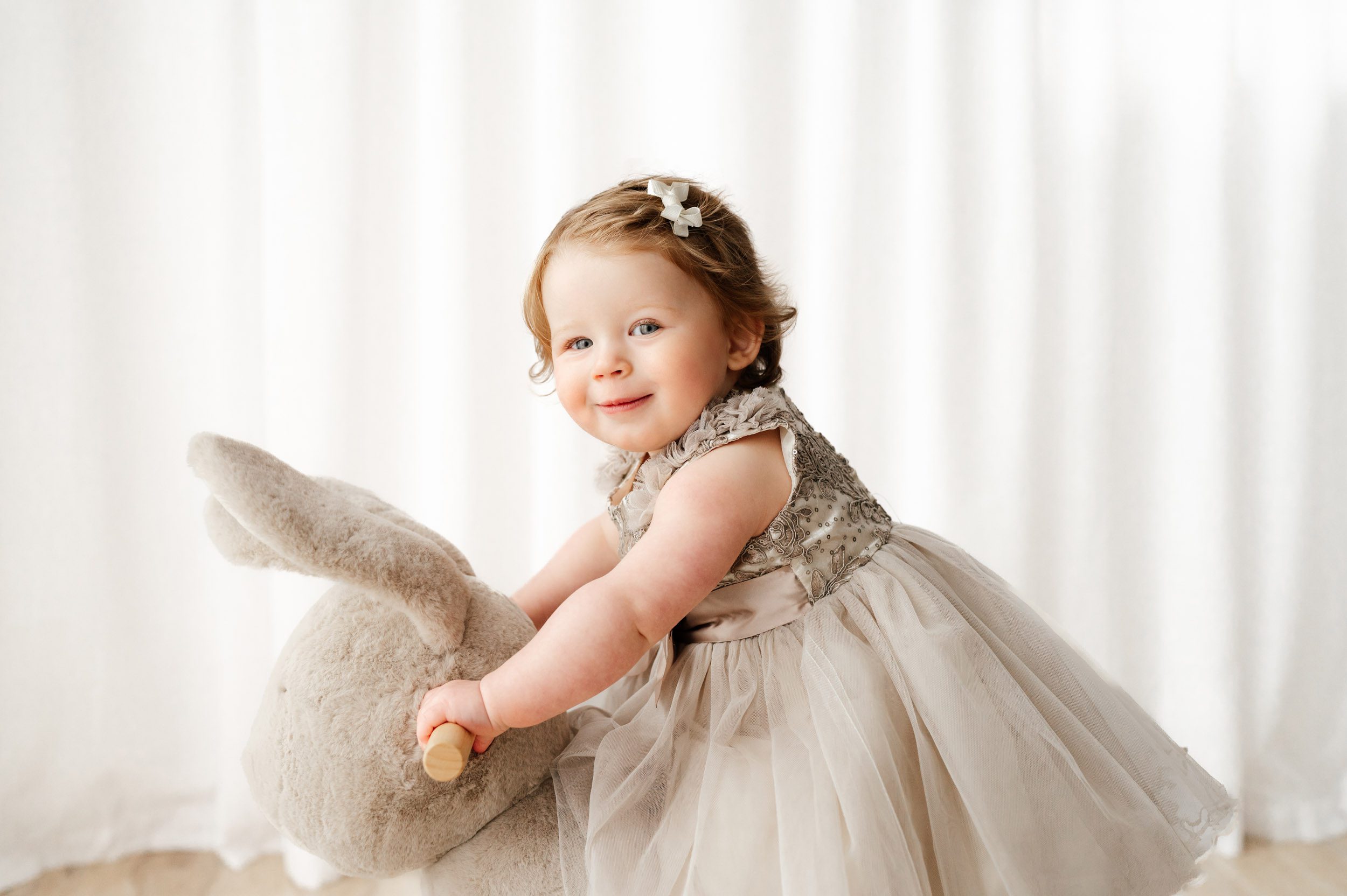 a young girl riding on a bunny rocker toy and looking as she looks at the camera and smiles during a 1st birthday photoshoot