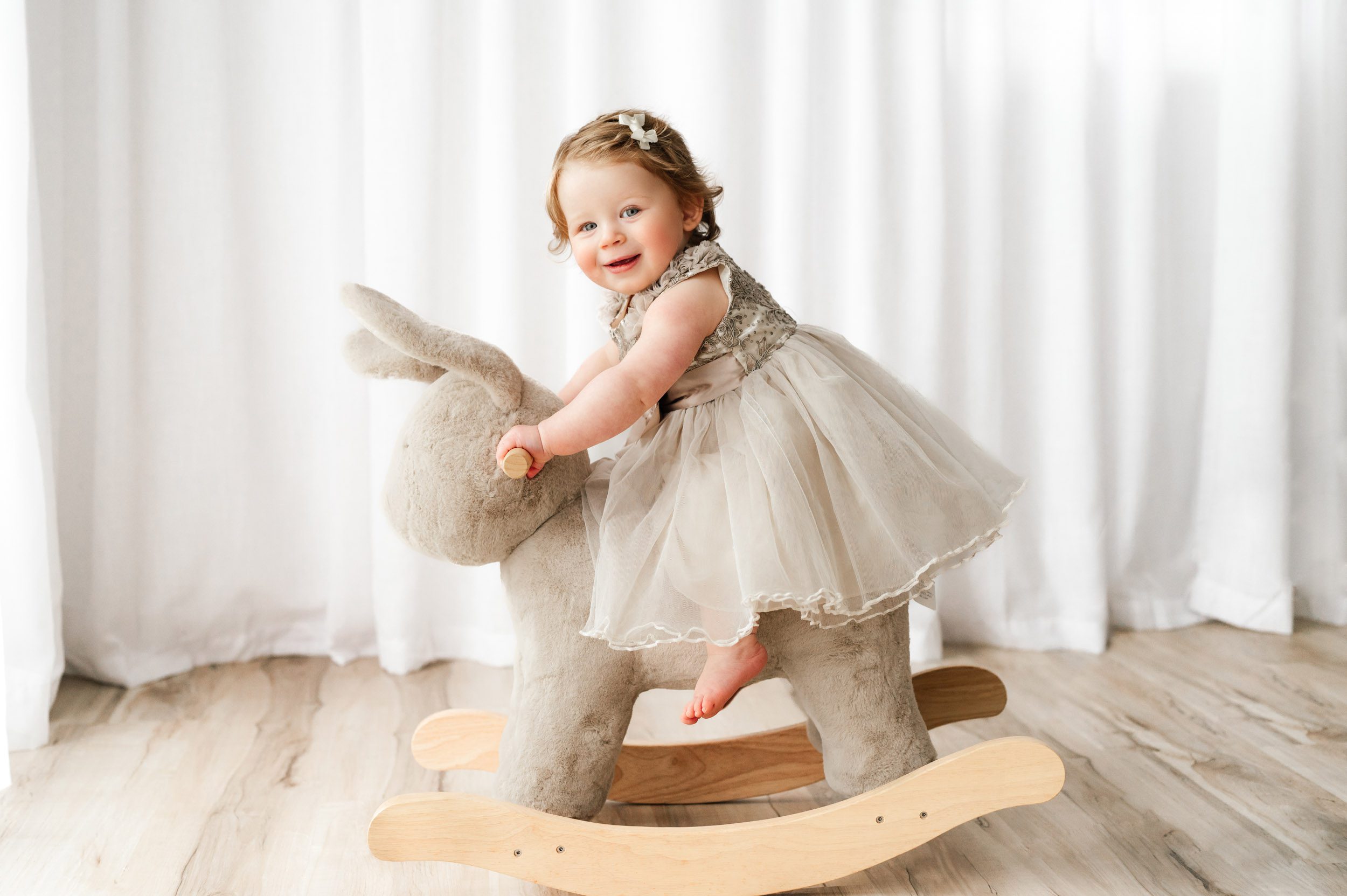 a young girl riding on a bunny rocker toy and laughing as she smiles at the camera during a toddler milestone photo session