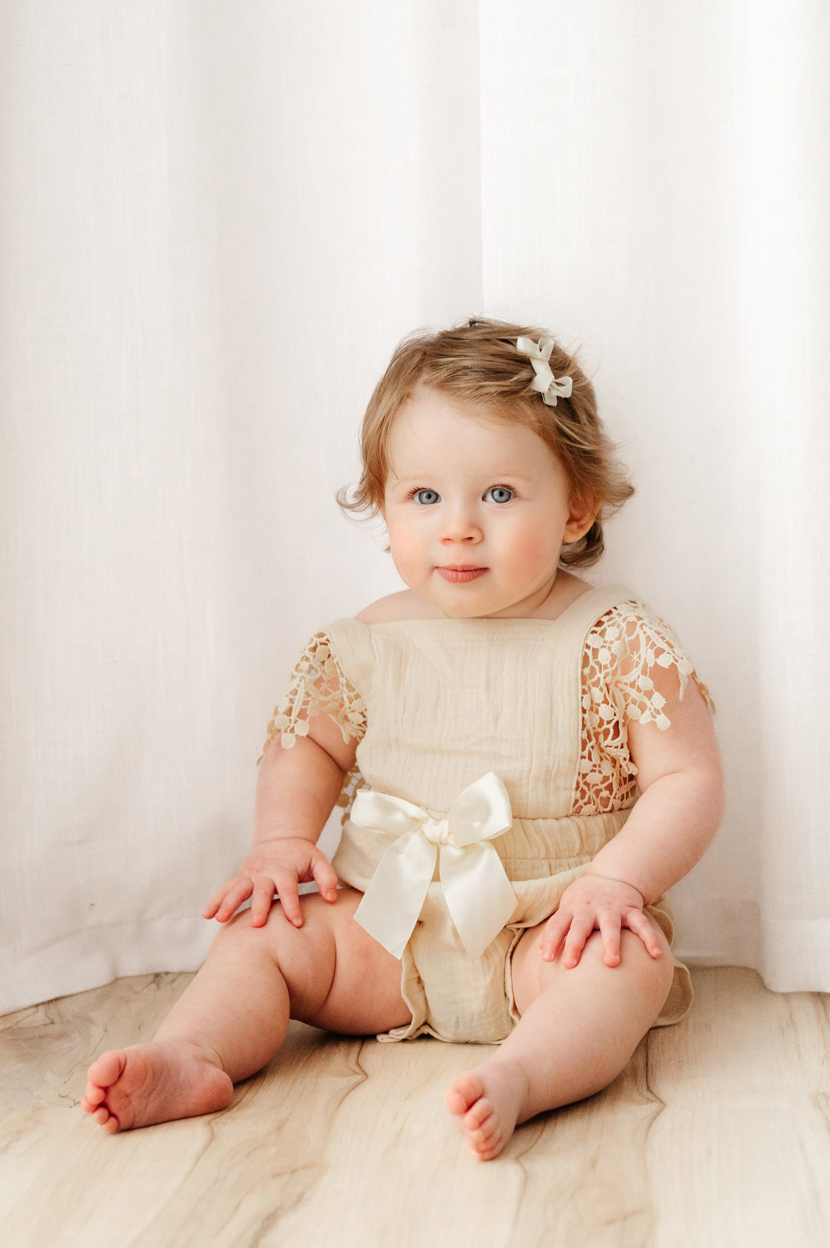 a young girl sitting in front of a white curtain with her hands on her knees and looking up above the camera at her parents during a 1st birthday photos session