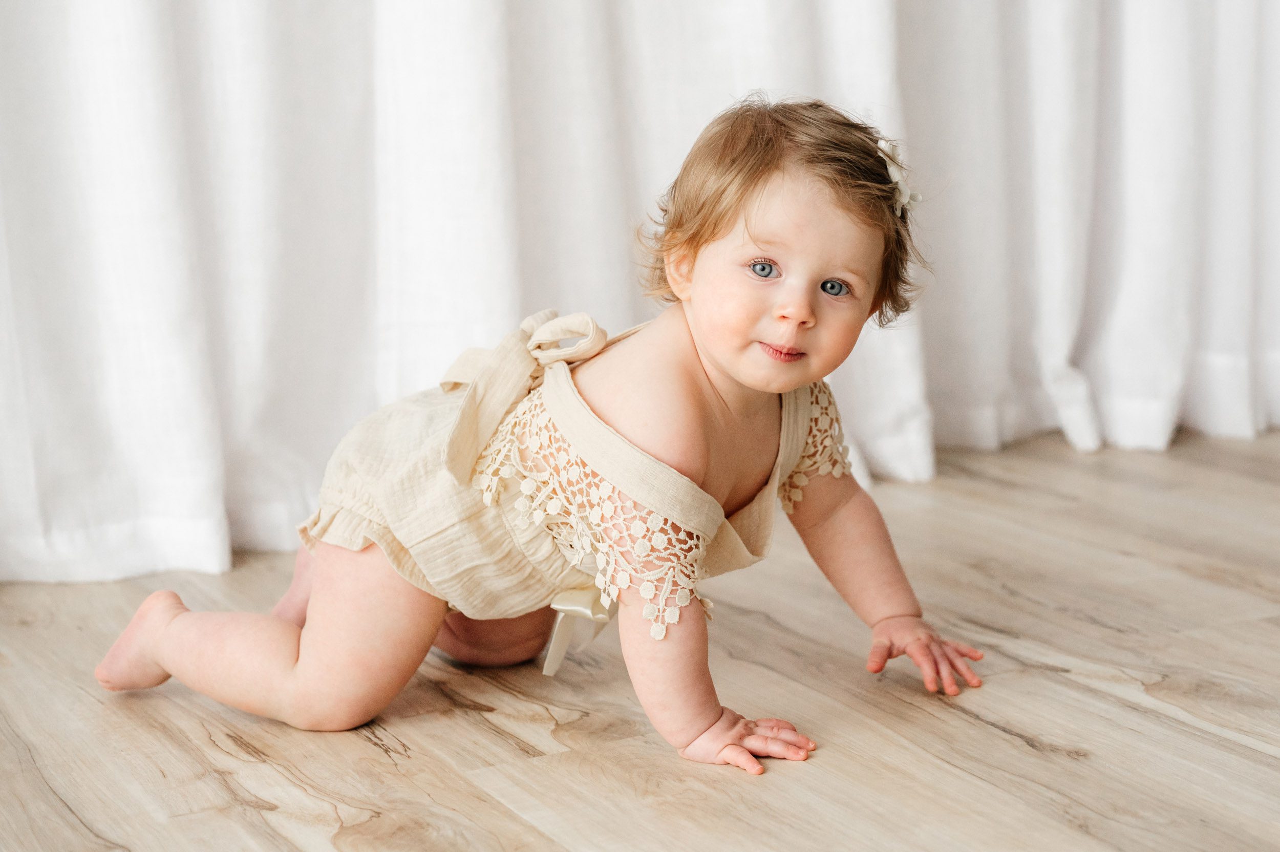 a young girl in a cream romper crawling across the floor with a mischievous look on her face during a 1st birthday photoshoot