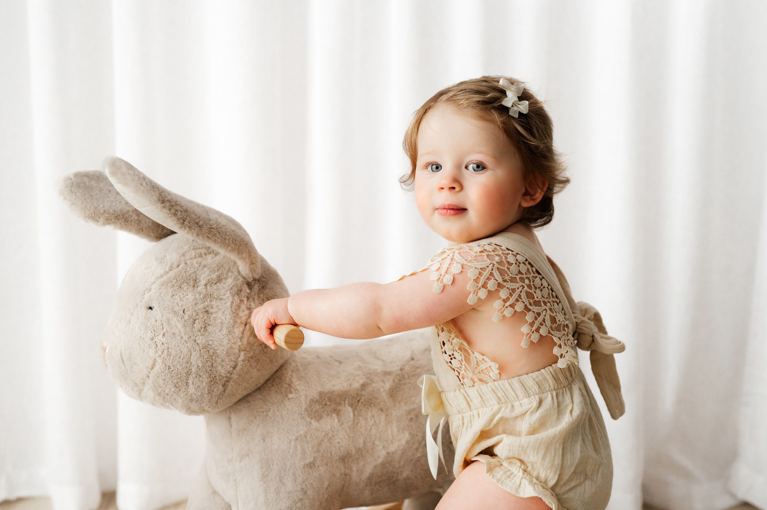 a little girl standing and holding onto a bunny rocker toy as she looks over her shoulder at the camera during a milestone photo session