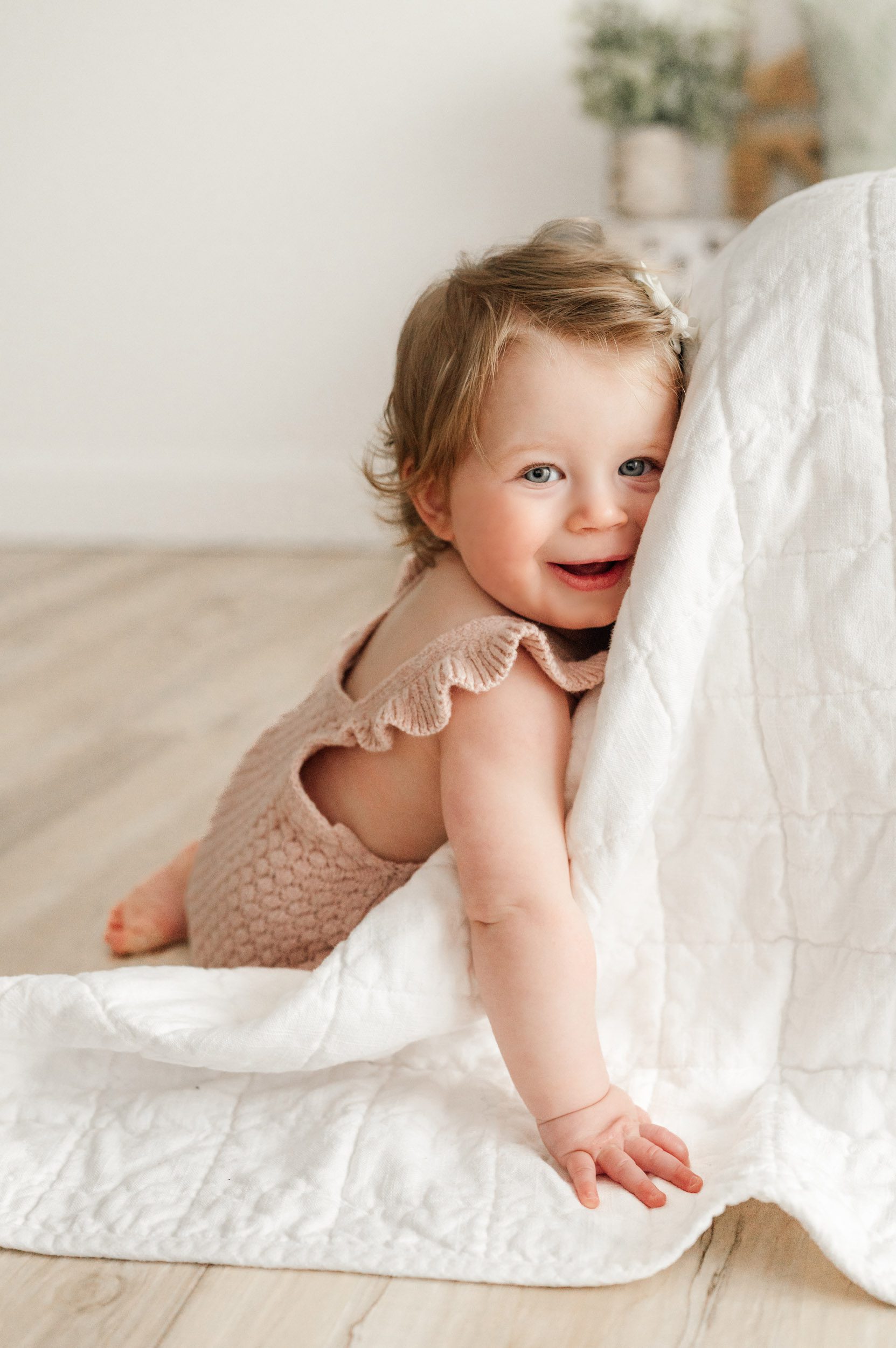 a little girl sitting next to a bed and resting her head against the comforter as she looks at the camera with a big smile on her face during a 1st birthday milestone photo session