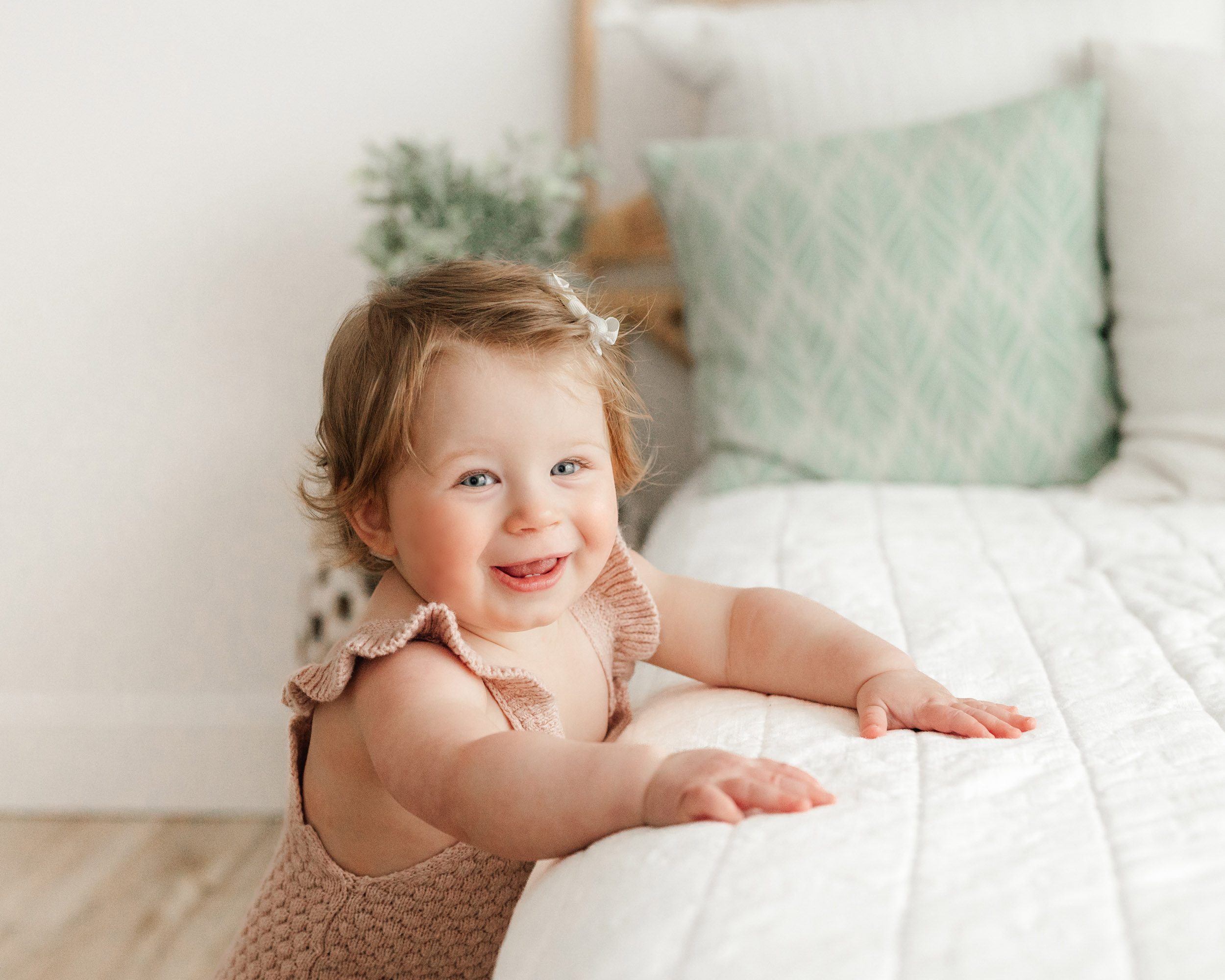 a young girl wearing a knit pink romper standing next to a bed and resting her arms on the bed as she looks at the camera with a huge smile on her face during a 1st birthday photo session