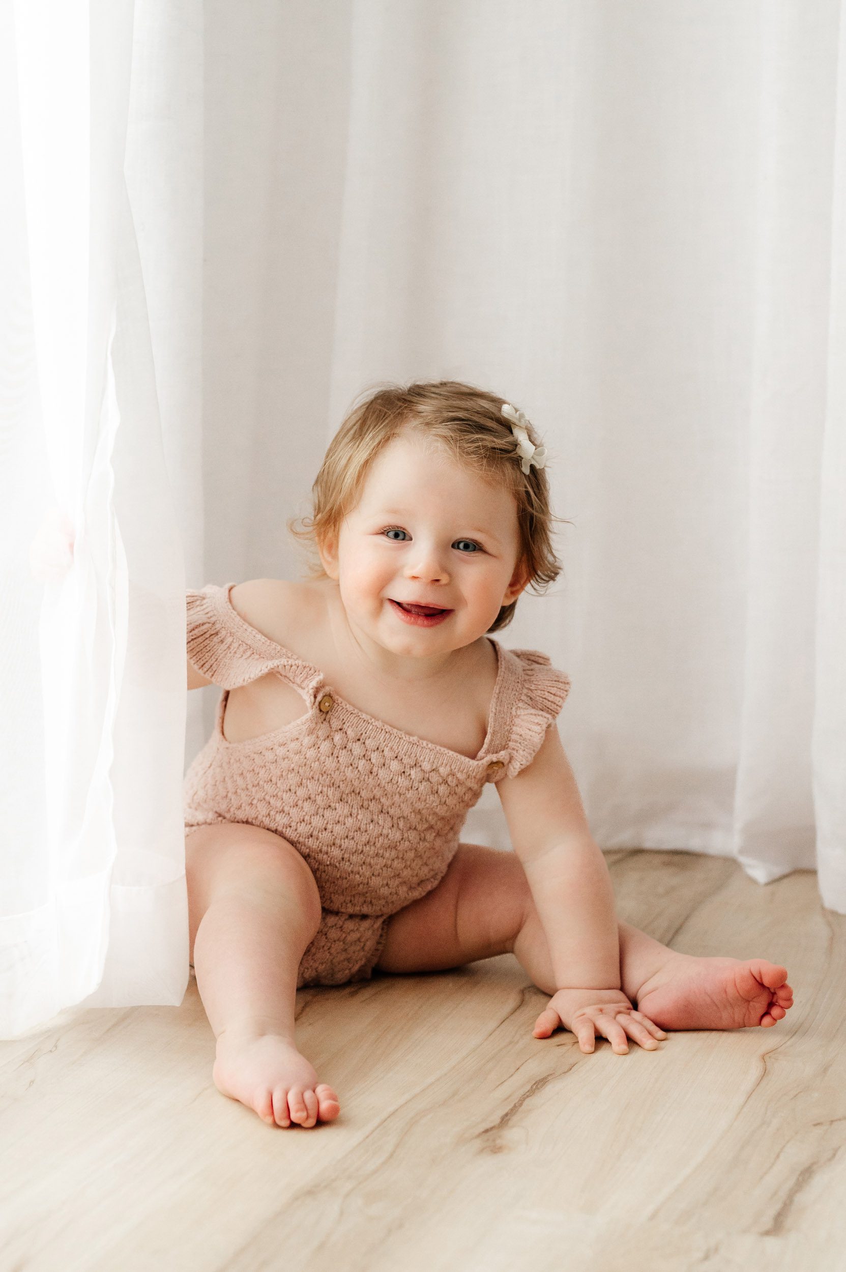 a young girl sitting in front of a white curtain and leaning forward and smiling at the camera during a milestone photoshoot