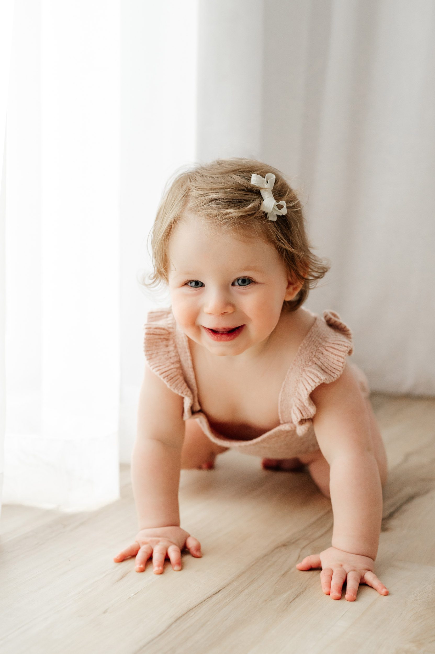 a young girl crawling on the floor next to window covered in sheer curtains and looking at the camera with a huge smile on her face during a milestone photoshoot
