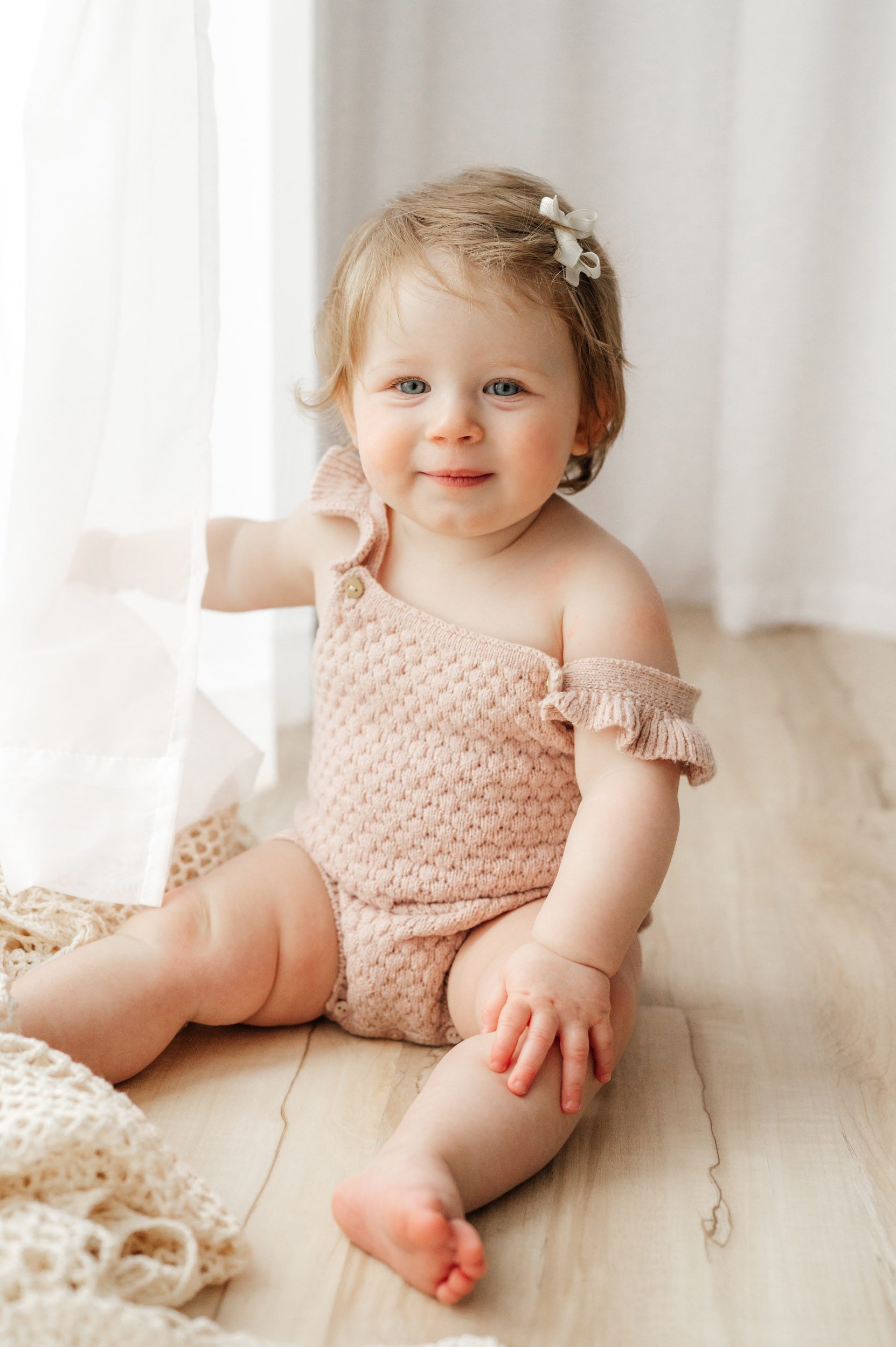 a young girl sitting on the floor and reaching out to touch the curtains that are covering the window next to her during a 1st birthday photos session