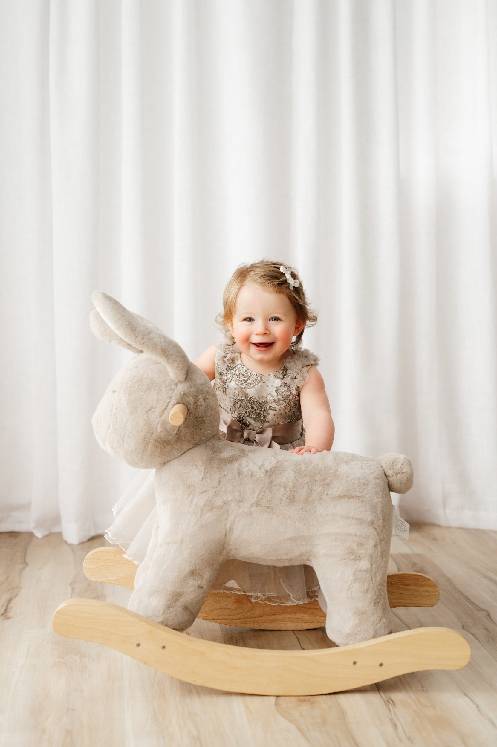 a young girl standing behind a bunny rocker toy and looking toward the camera and laughing as she rocks the bunny during a 1st birthday photos session