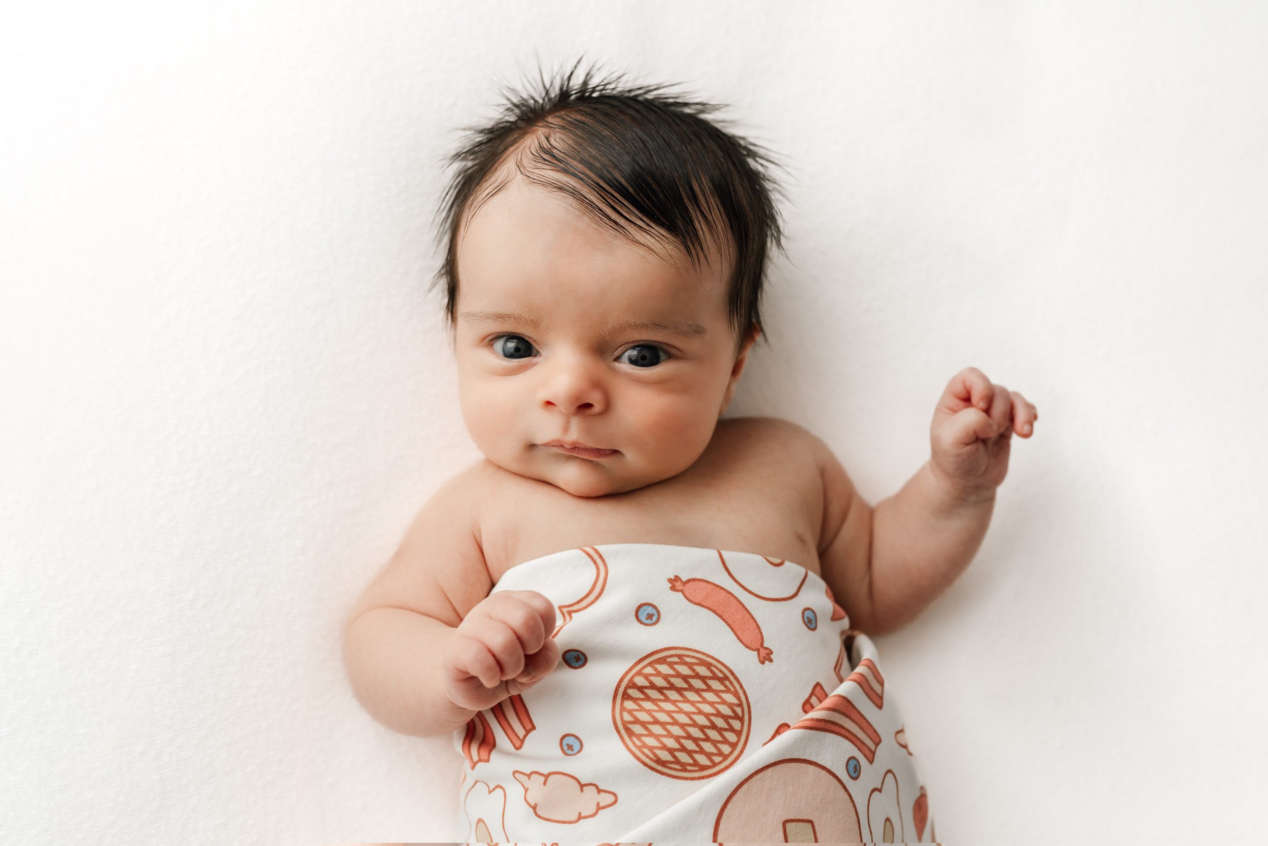 an older baby boy wrapped in a colorful swaddle laying on a white backdrop and looking directly up at the camera during a newborn photos session