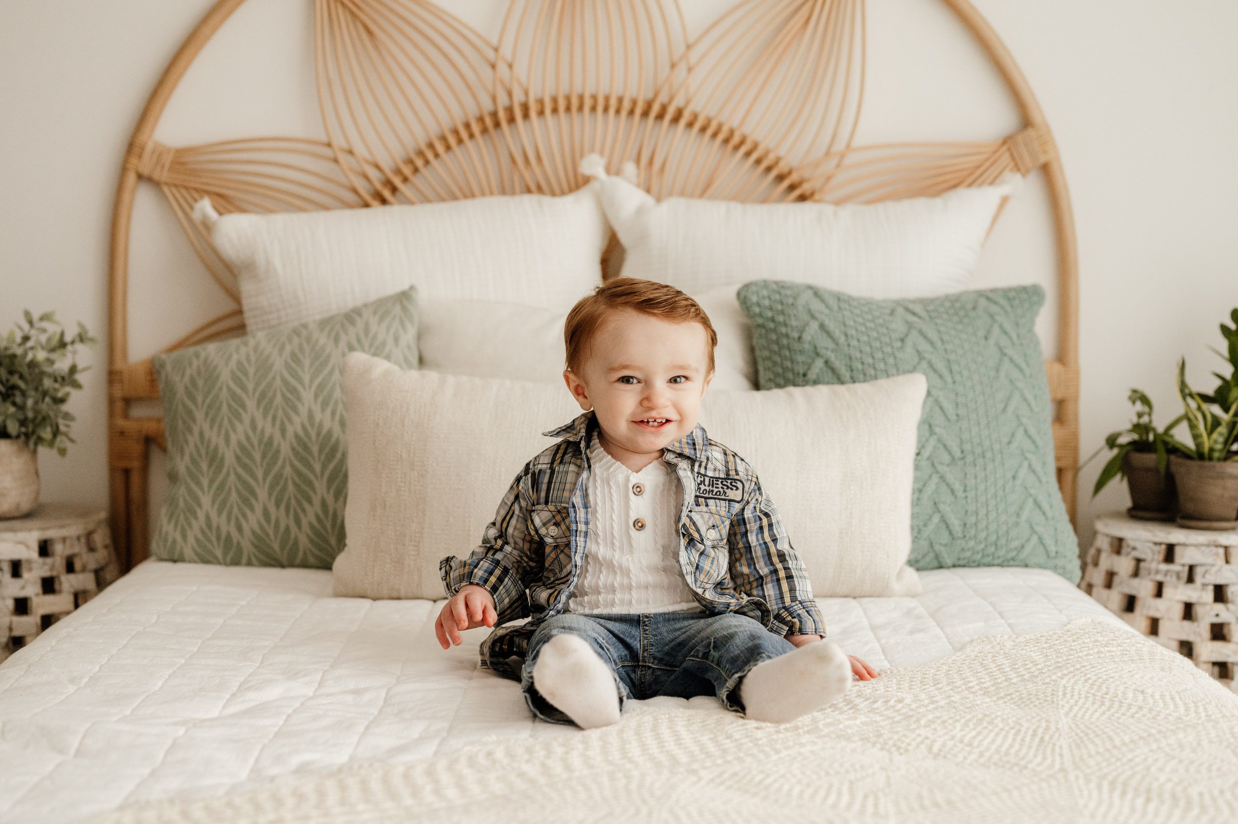 a young boy sitting on a bed and looking right at the camera with a huge smile on his face during a 1st birthday photo session