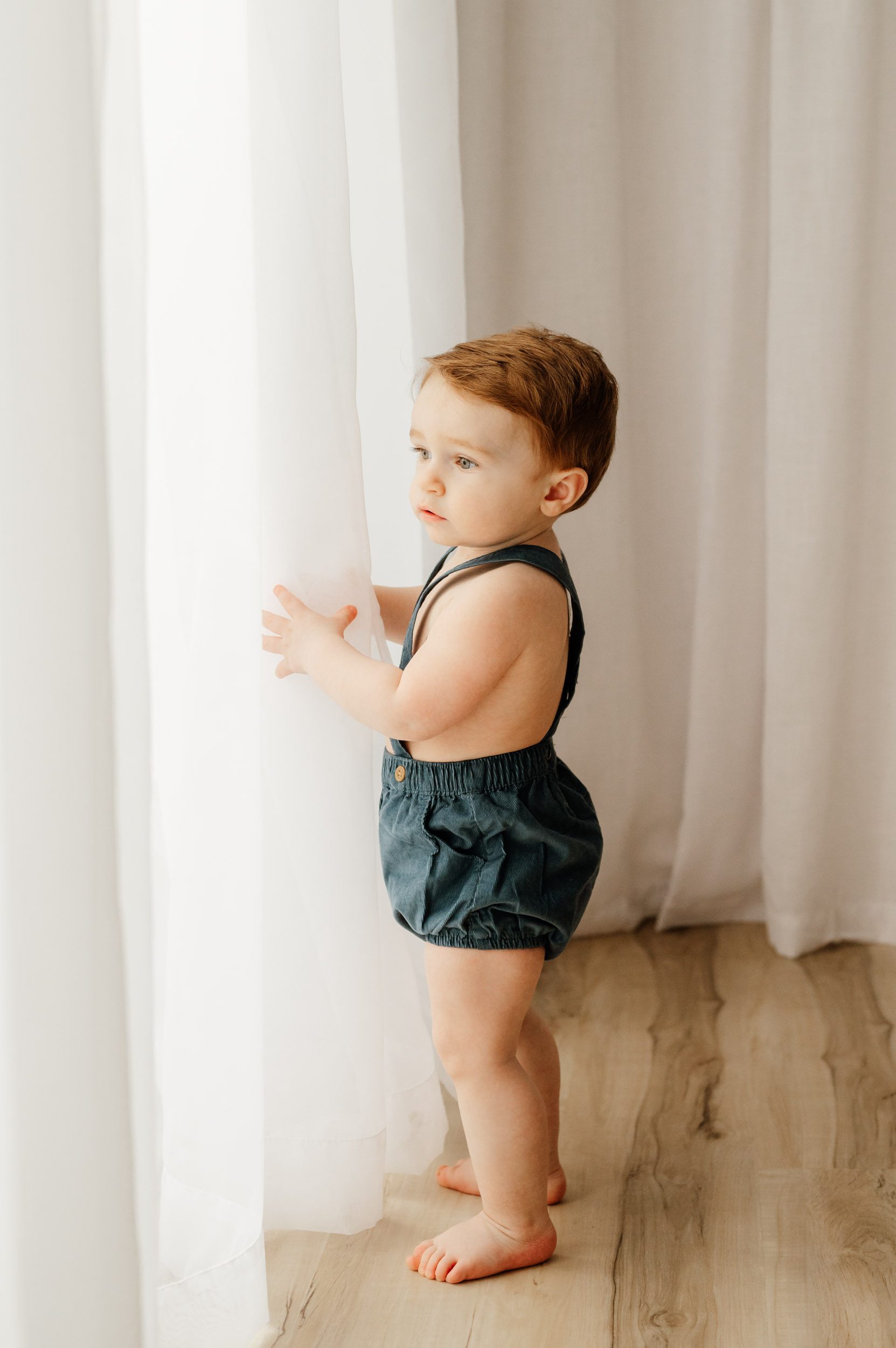 a young boy standing next to a window and reaching out to touch the sheer curtains and he gazes outside during a toddler milestone photography session