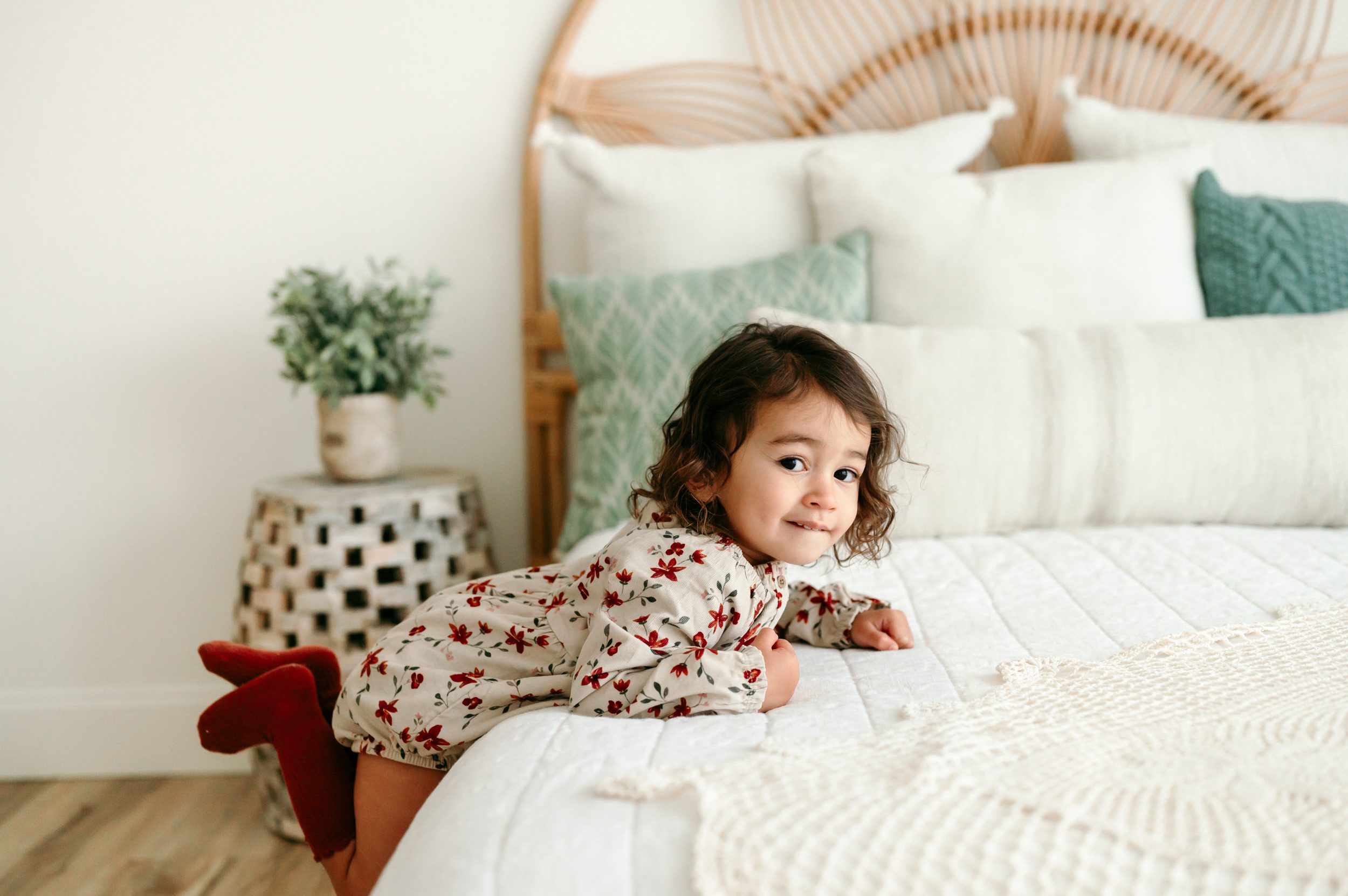 a young girl climbing up onto a bed and lifting her feet in the air as she looks at the camera and bites her lower lip during a toddler milestone photography session