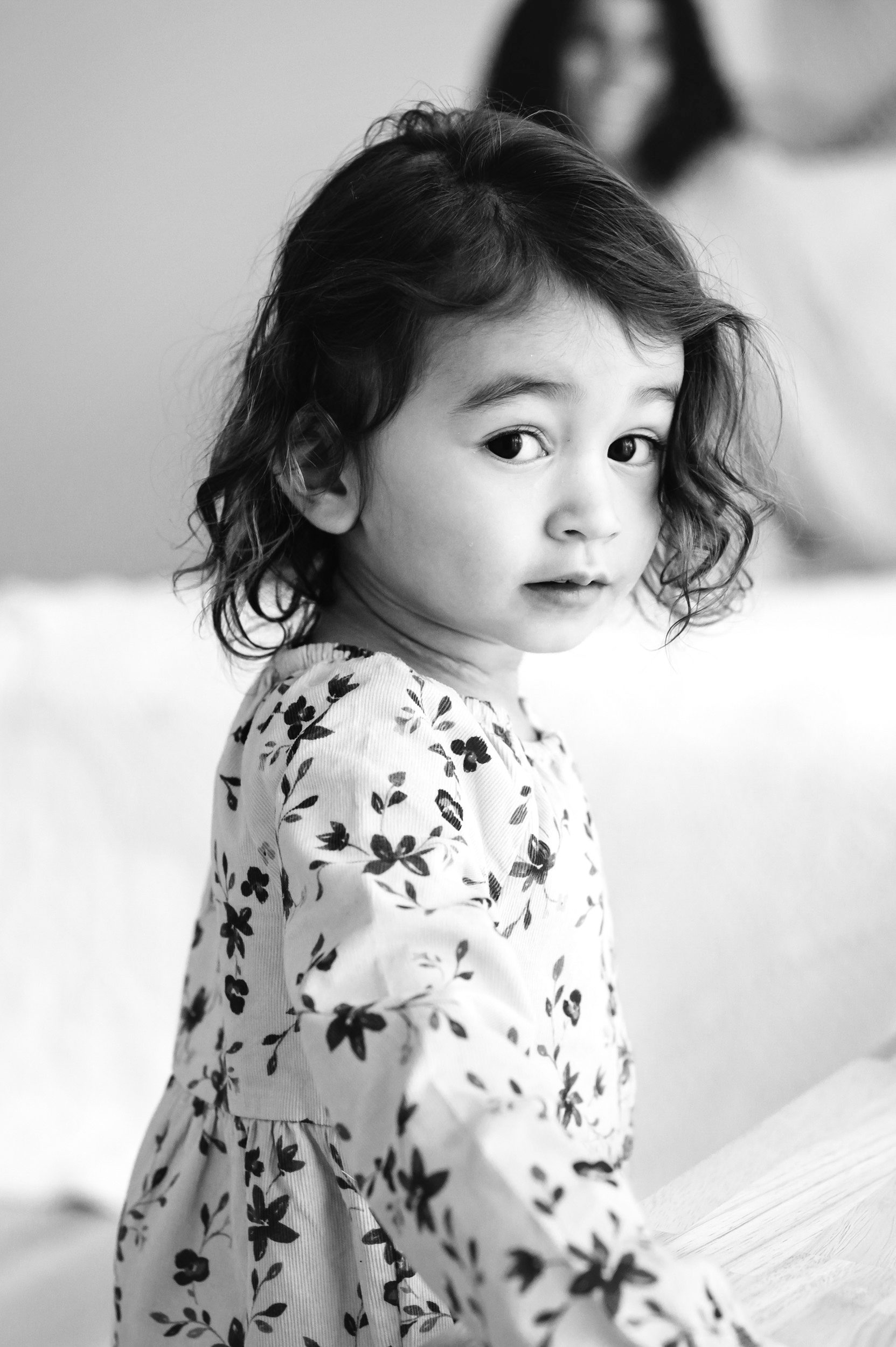 a black and white picture of a young girl looking straight at the camera with her mom sitting on the bed in the background smiling at her during a toddler milestone photo session