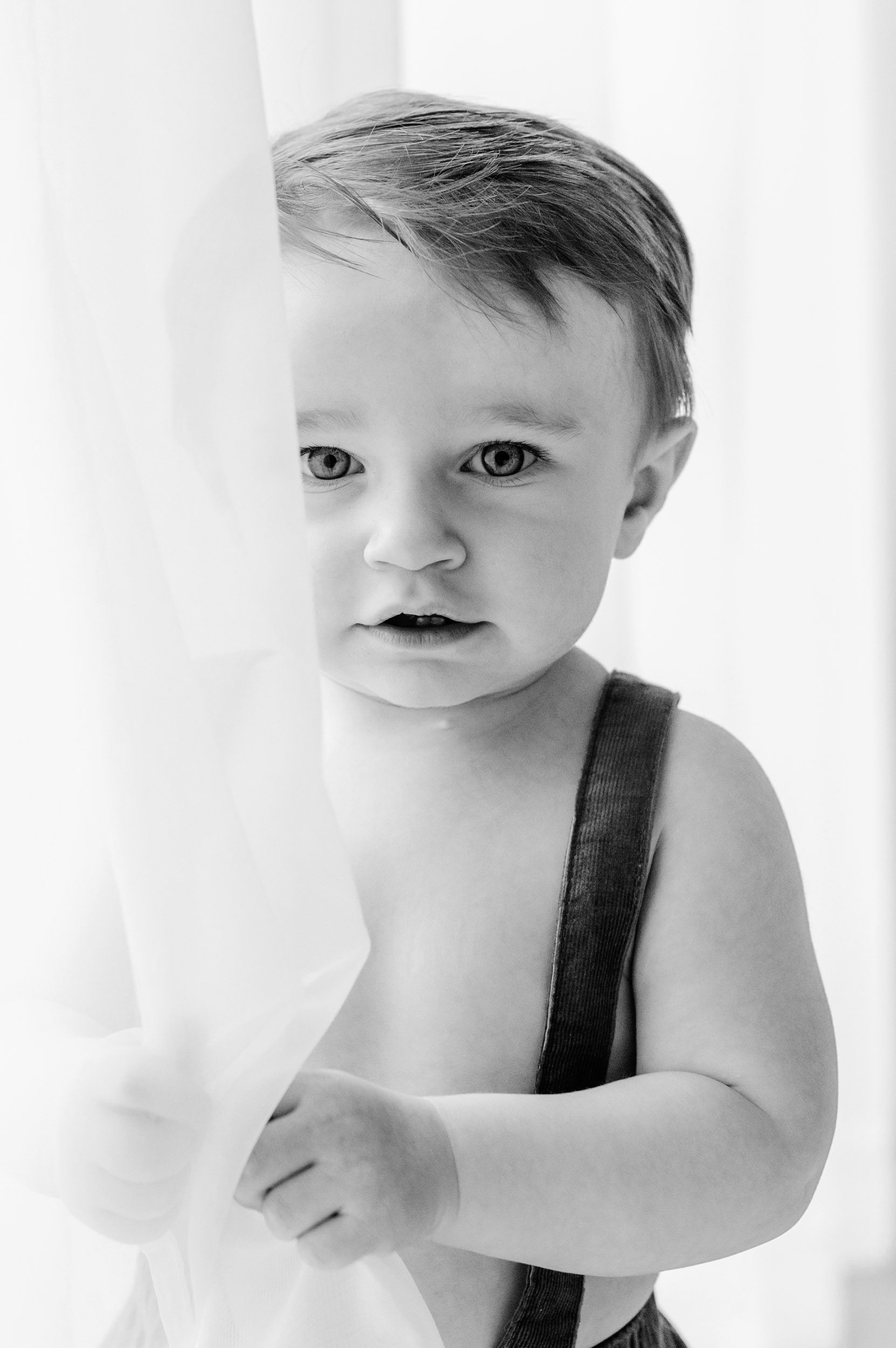 a black and white picture of a young boy holding a sheer curtain in his hand and peeking out from behind it looking directly at the camera during a toddler milestone photoshoot
