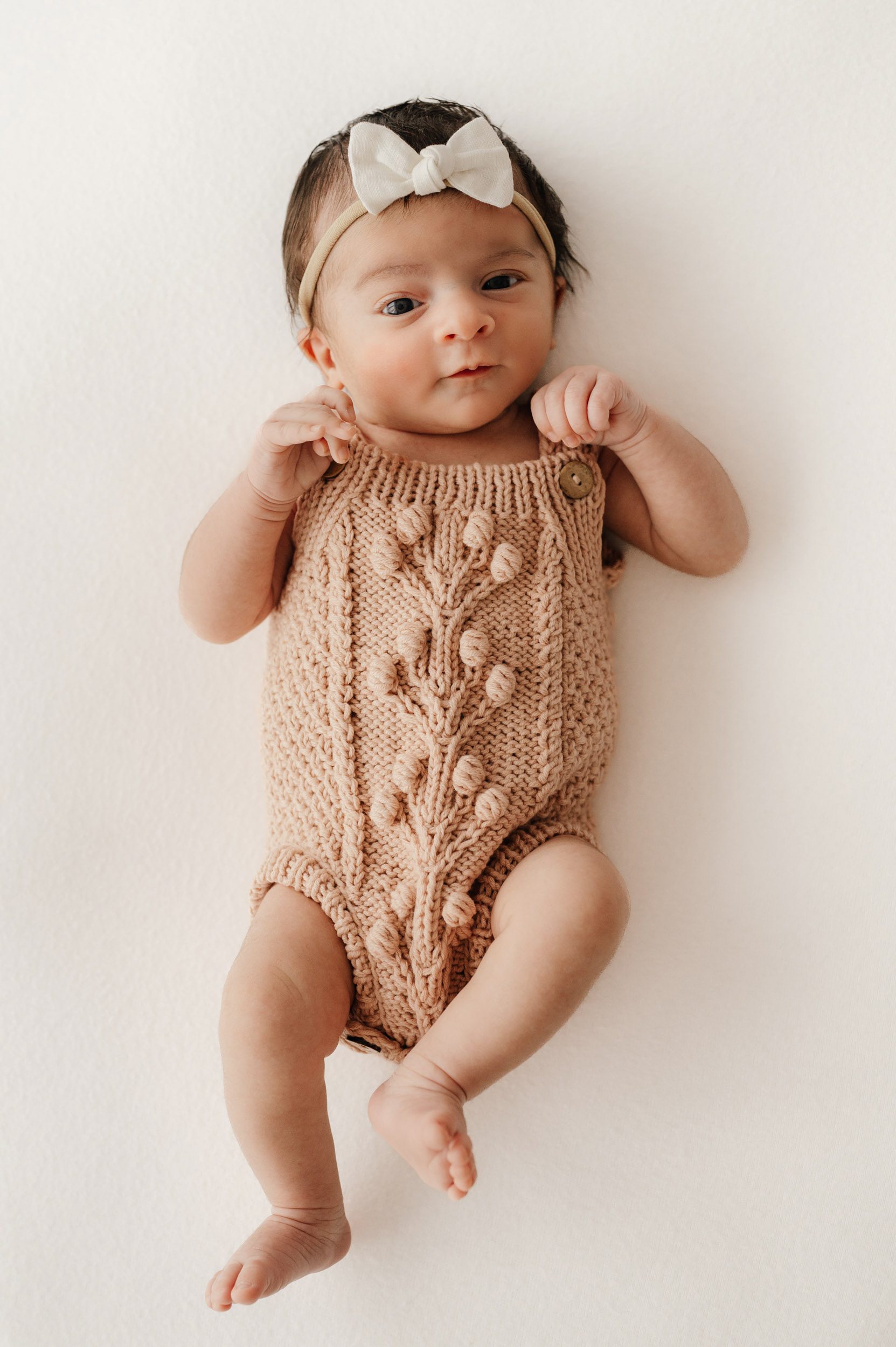 a baby girl wearing a light pink knit onesie and white bow headband laying on her back on a white backdrop looking directly up at the camera during a natural light newborn photoshoot