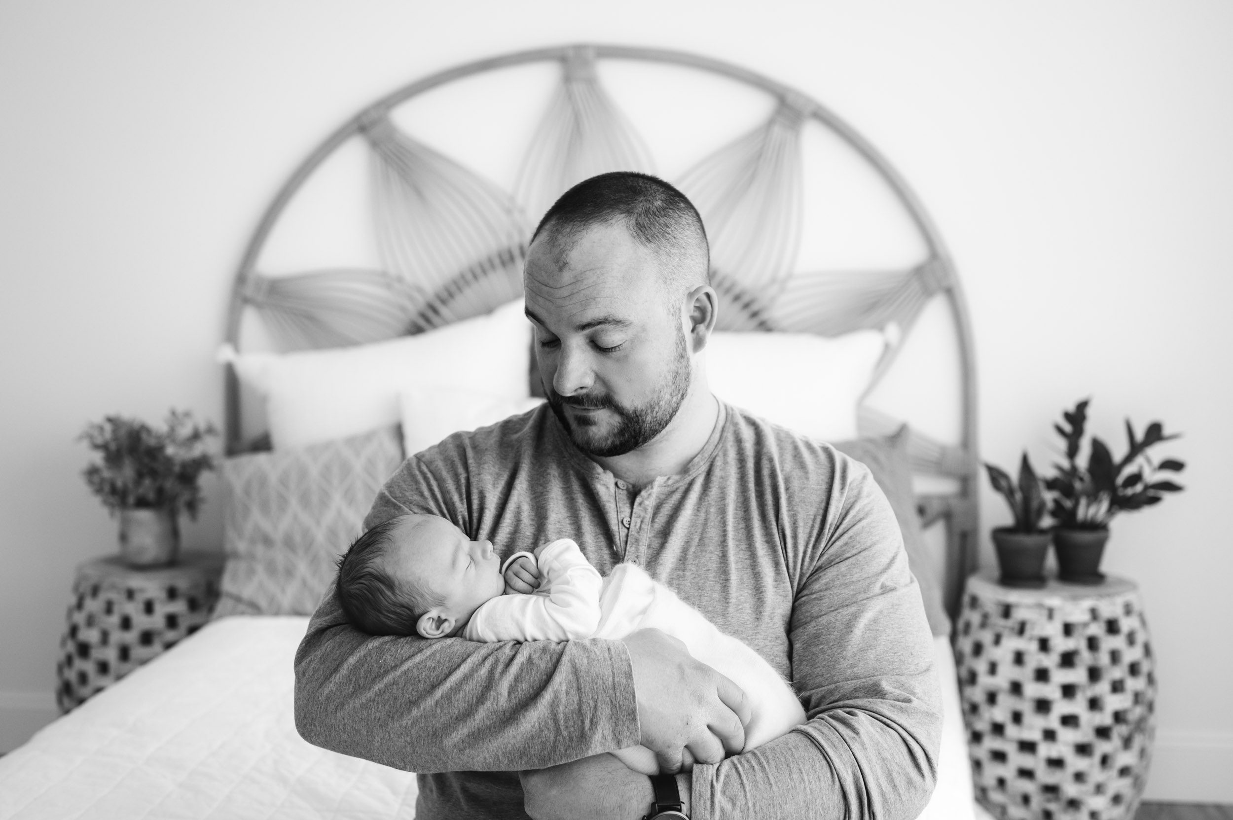 a black and white picture of a dad sitting on a bed and looking down at his newborn son cradled in his arms during a lifestyle newborn photo session