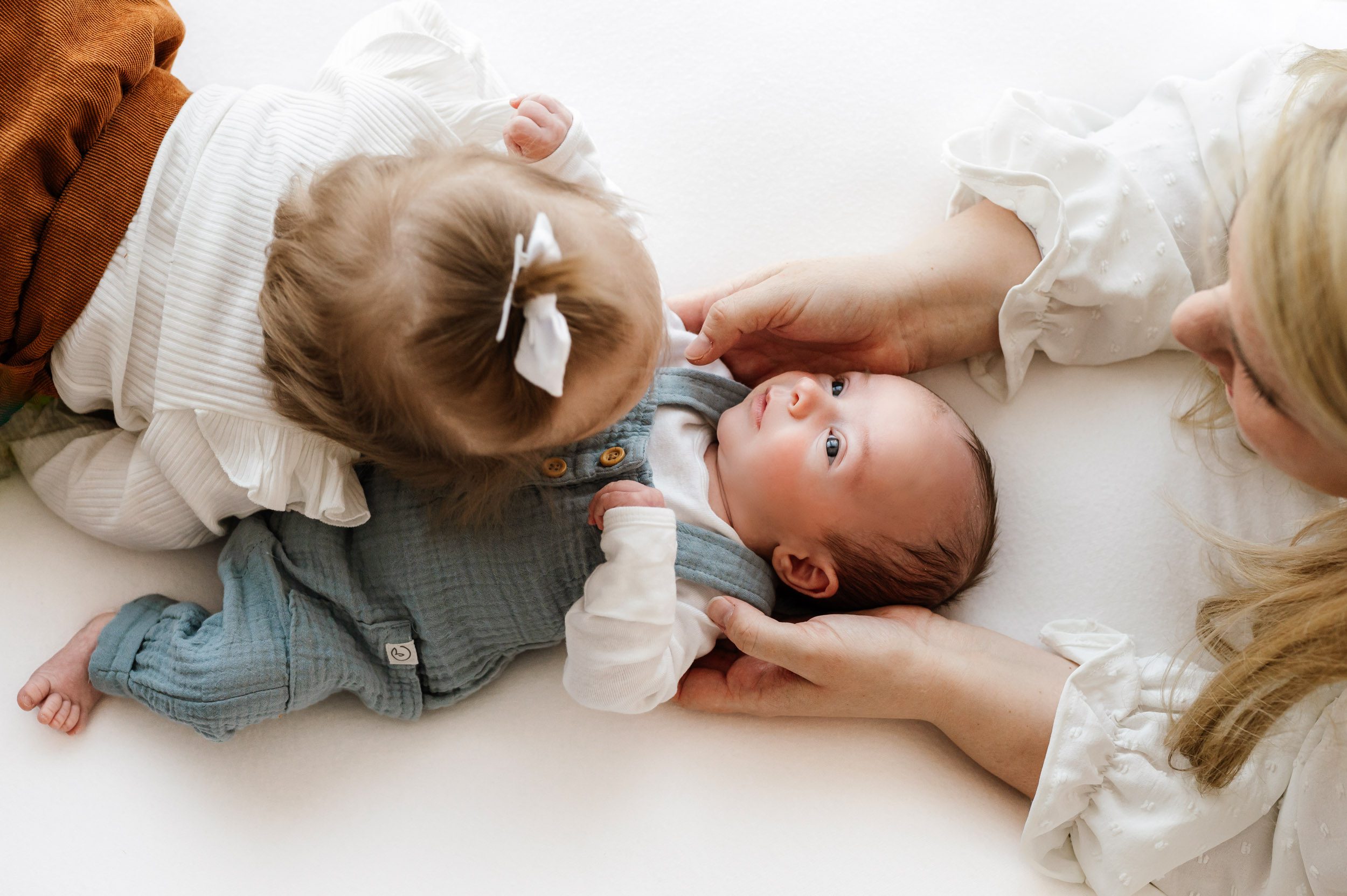 a picture taken from above of a baby boy wearing blue linen overalls laying on a white backdrop while is mom and big sister cuddle him as he looks toward the window light during a lifestyle newborn photoshoot
