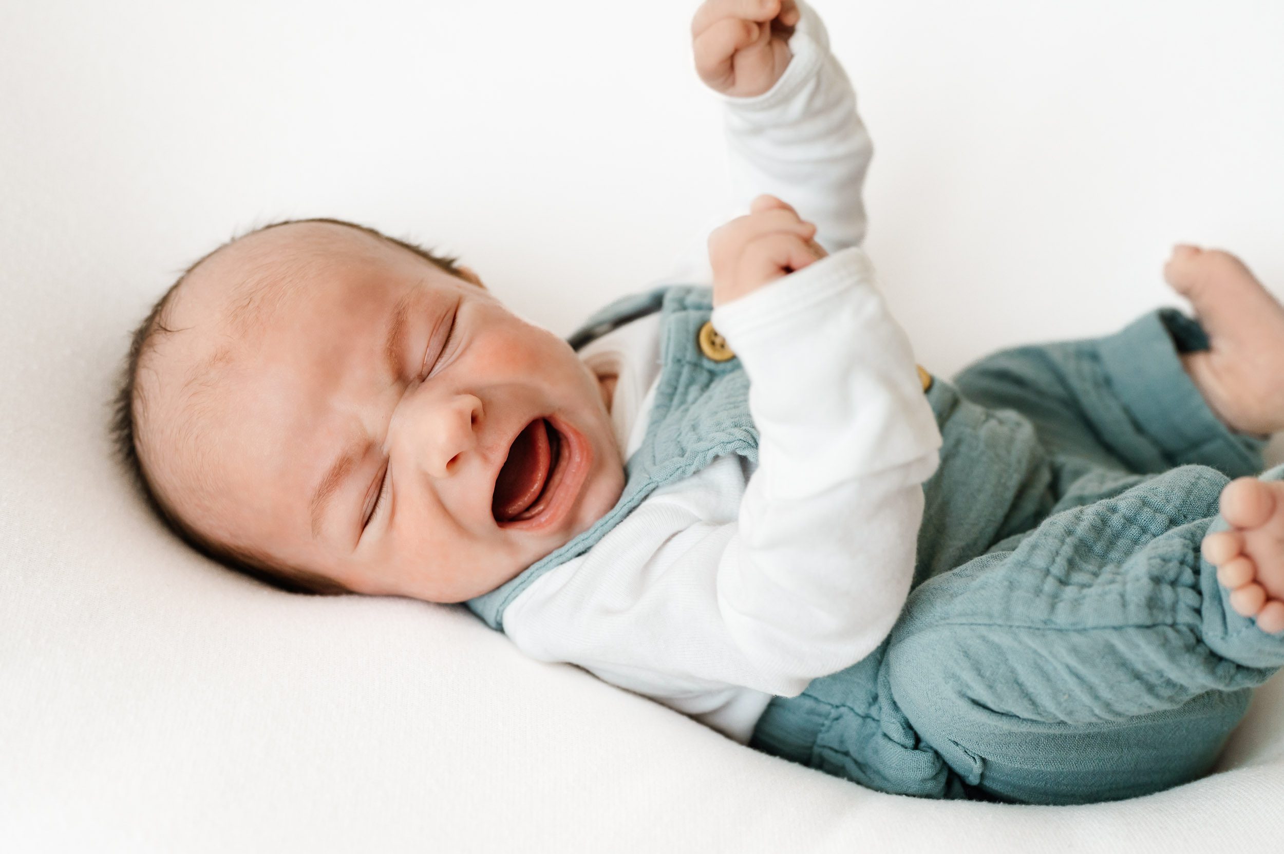 a baby boy wearing blue linen overalls and laying on a white backdrop lifting his hands and feet in the air as he cries during a lifestyle newborn photoshoot