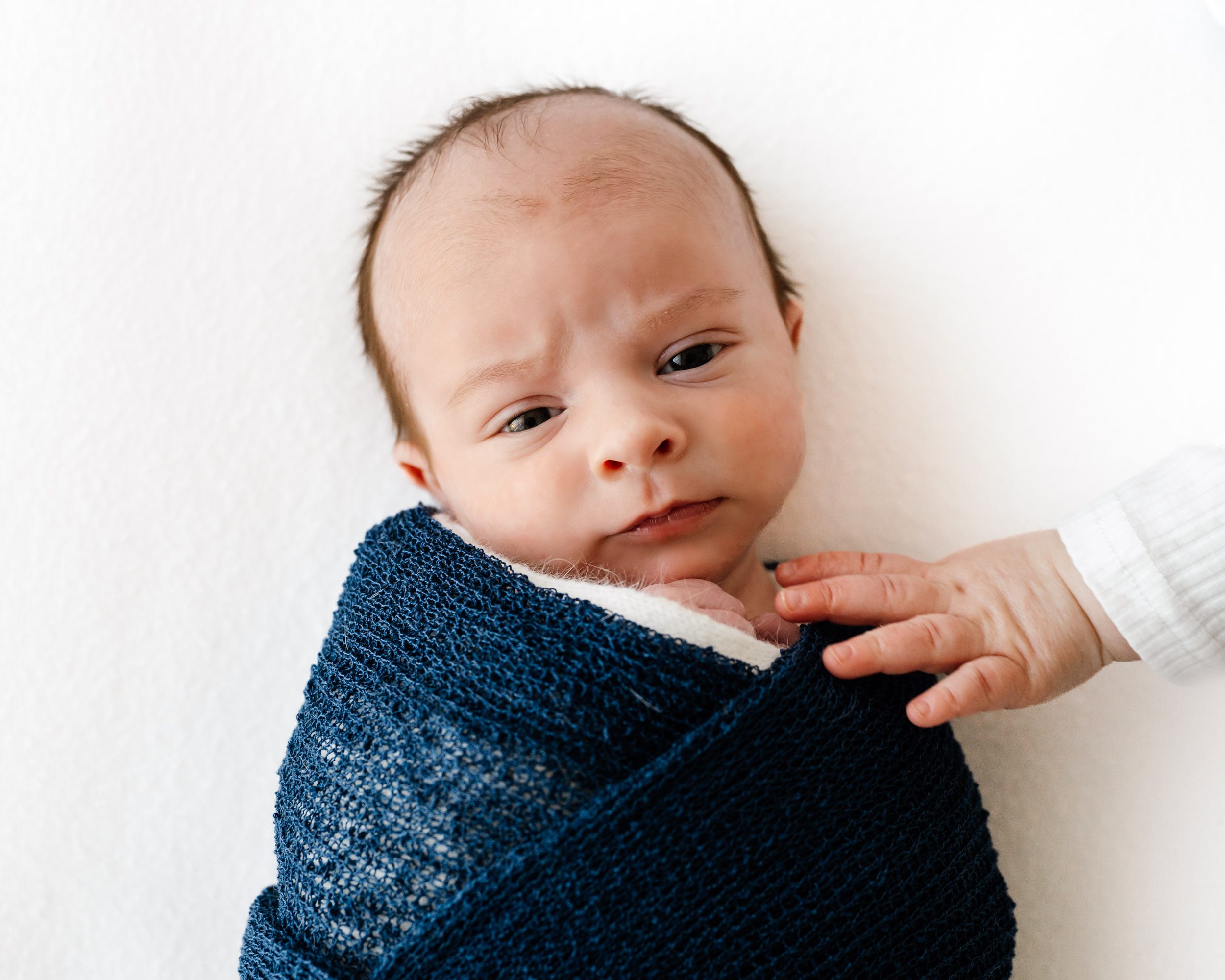 a baby boy wrapped in a blue swaddle blanket laying on a white backdrop and looking directly at the camera while his big sister reaches out to touch him with her hand during a lifestyle newborn photo session