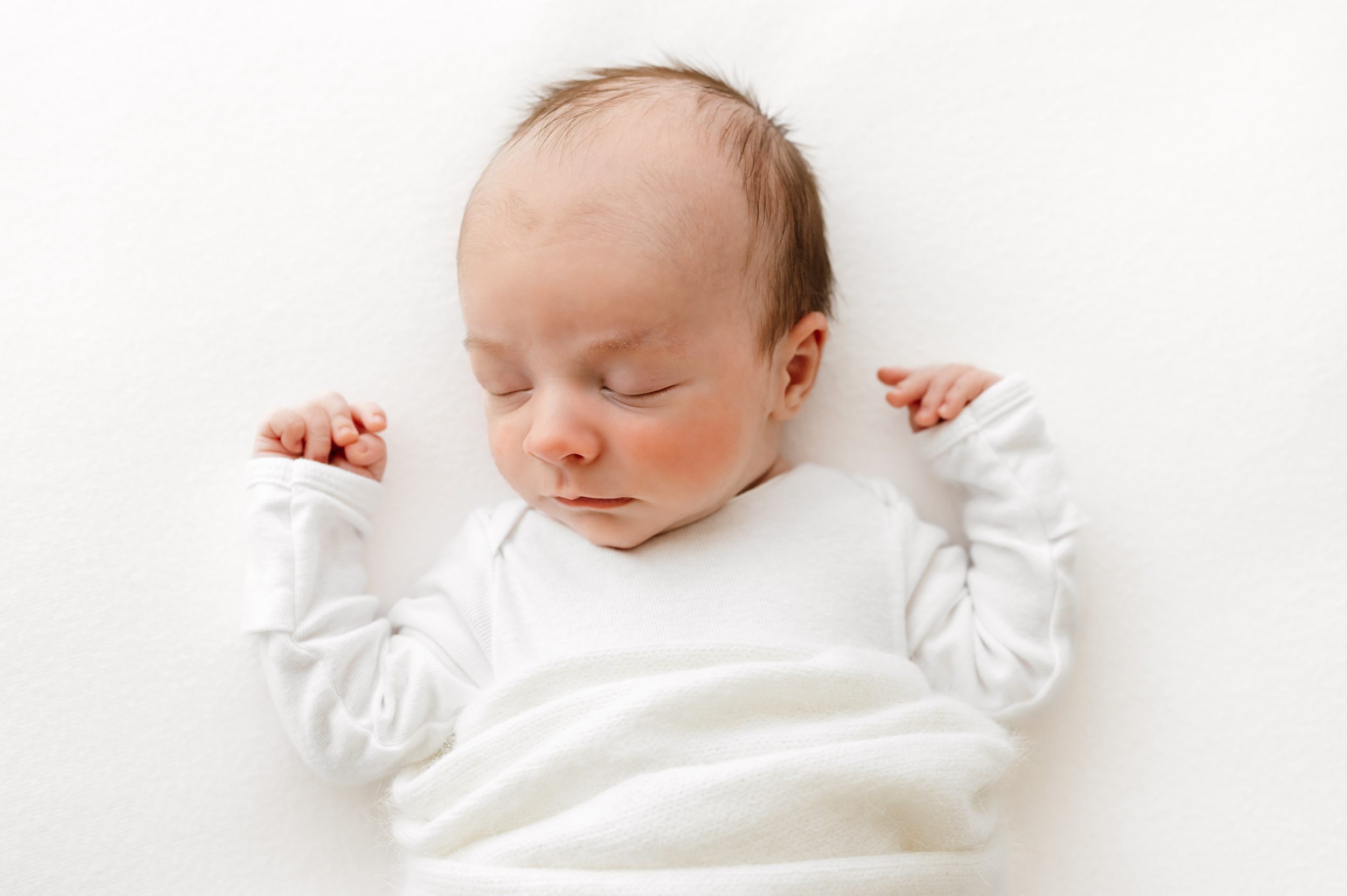 a baby boy wearing a long sleeved white onesie and sleeping on his back on a white backdrop during a lifestyle newborn photoshoot