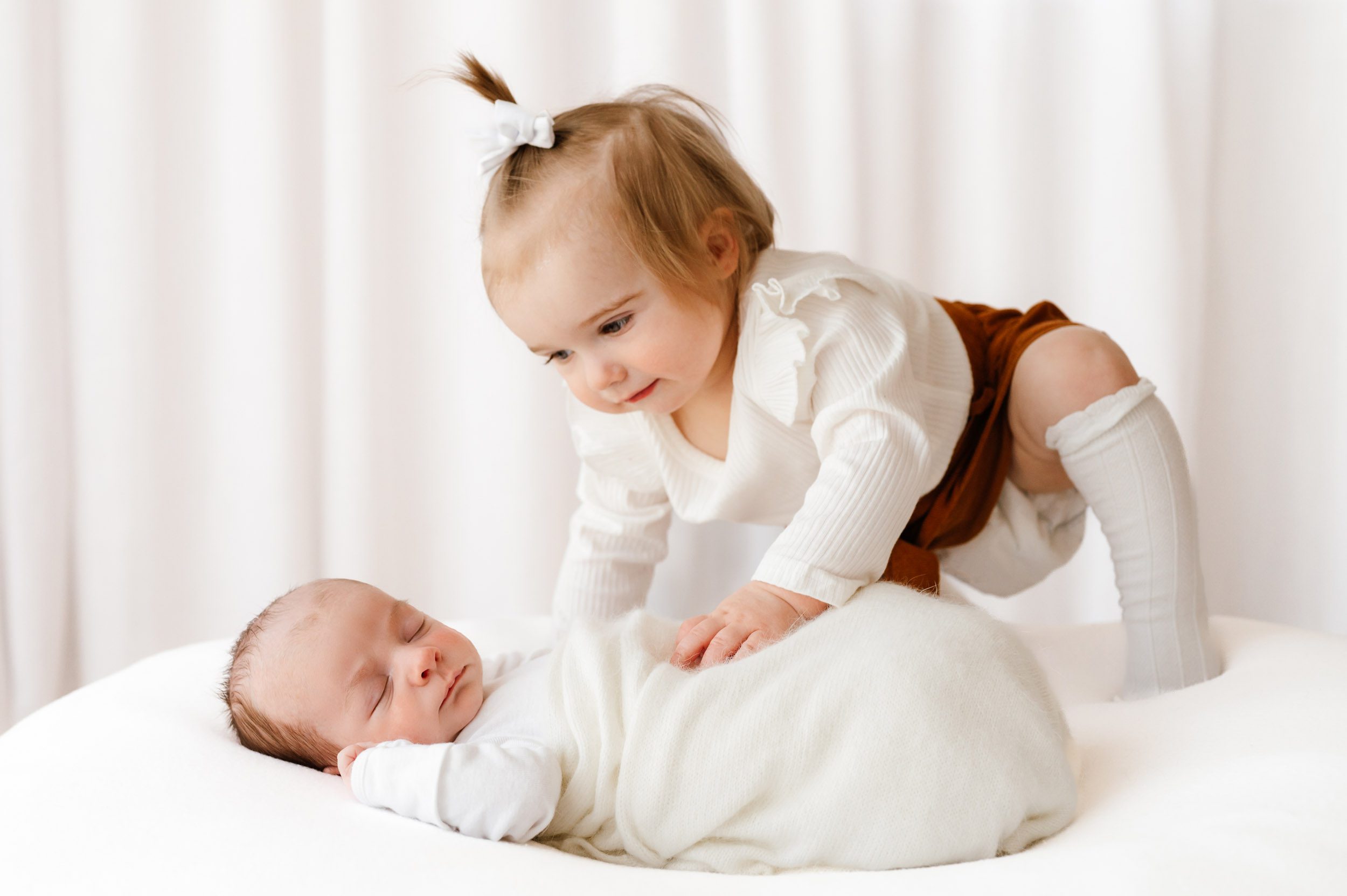 a baby boy wrapped in a white swaddle sleeping on a white bean bag while his big sister climbs up onto the bean bag and gently touches his belly with her hand during a lifestyle newborn photo session