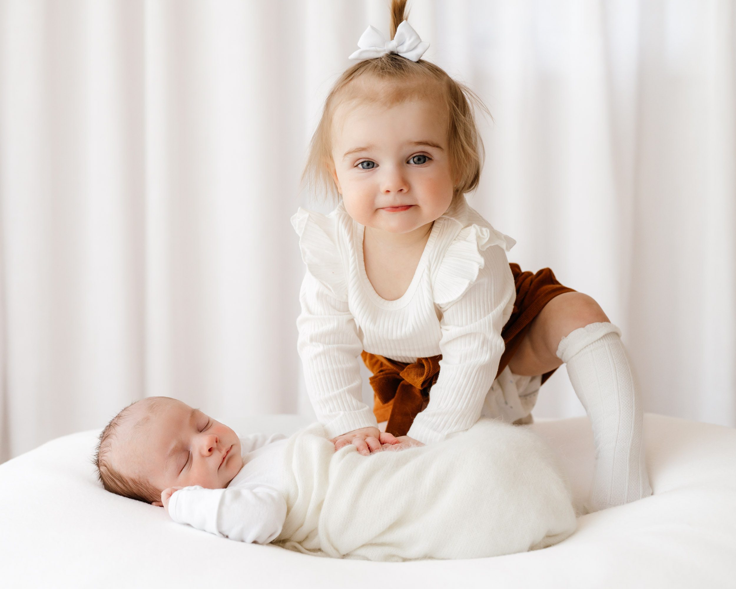a baby boy wrapped in a white swaddle blanket laying on a white bean bag while his big sister kneels above him looking directly at the camera with a hint of a smile on her face during a lifestyle newborn photoshoot
