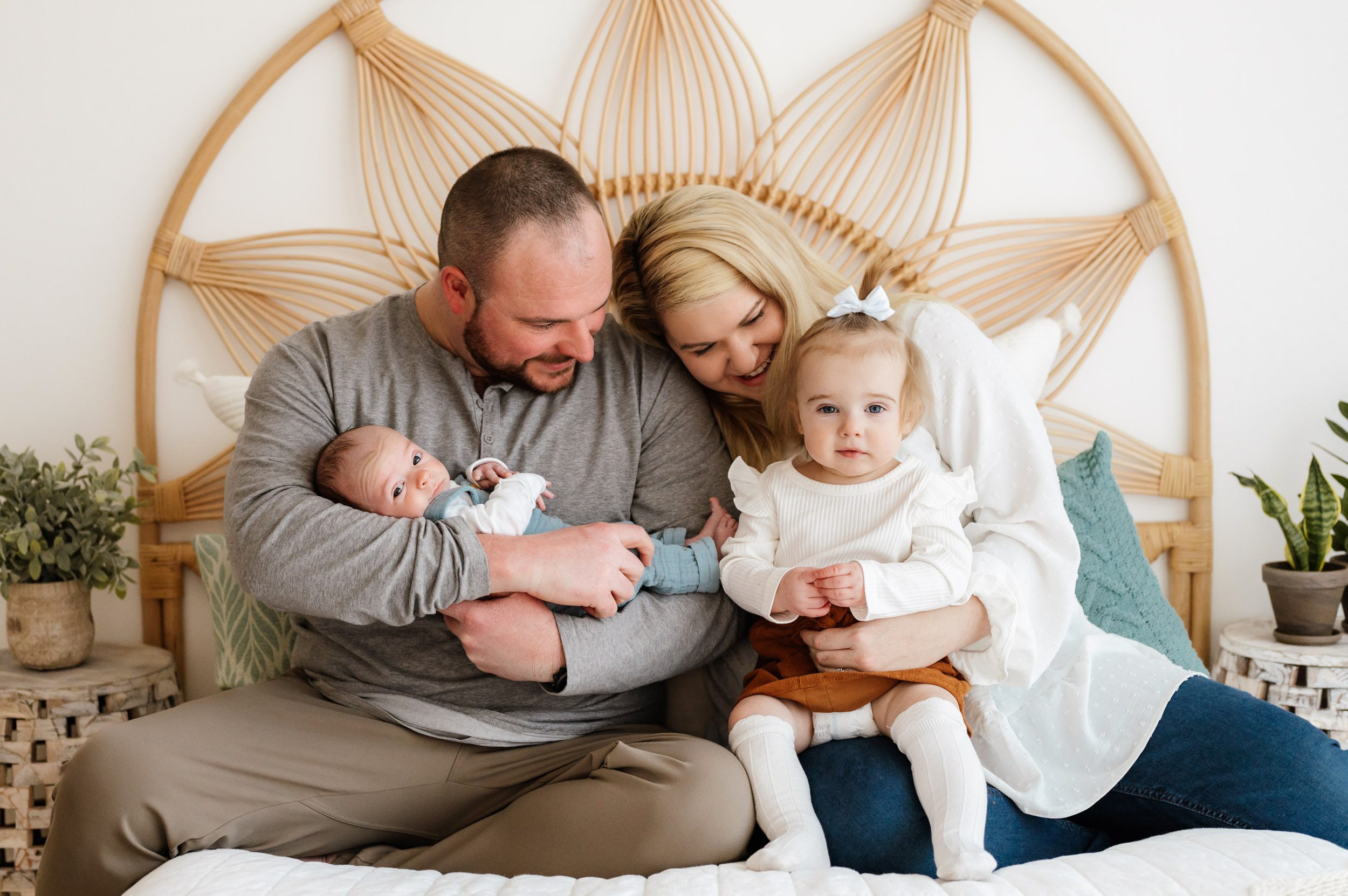 a new family of four sitting on a bed and snuggling with their new baby and toddler while both parents smile at the toddler and the baby and toddler look right at the camera during a newborn photoshoot