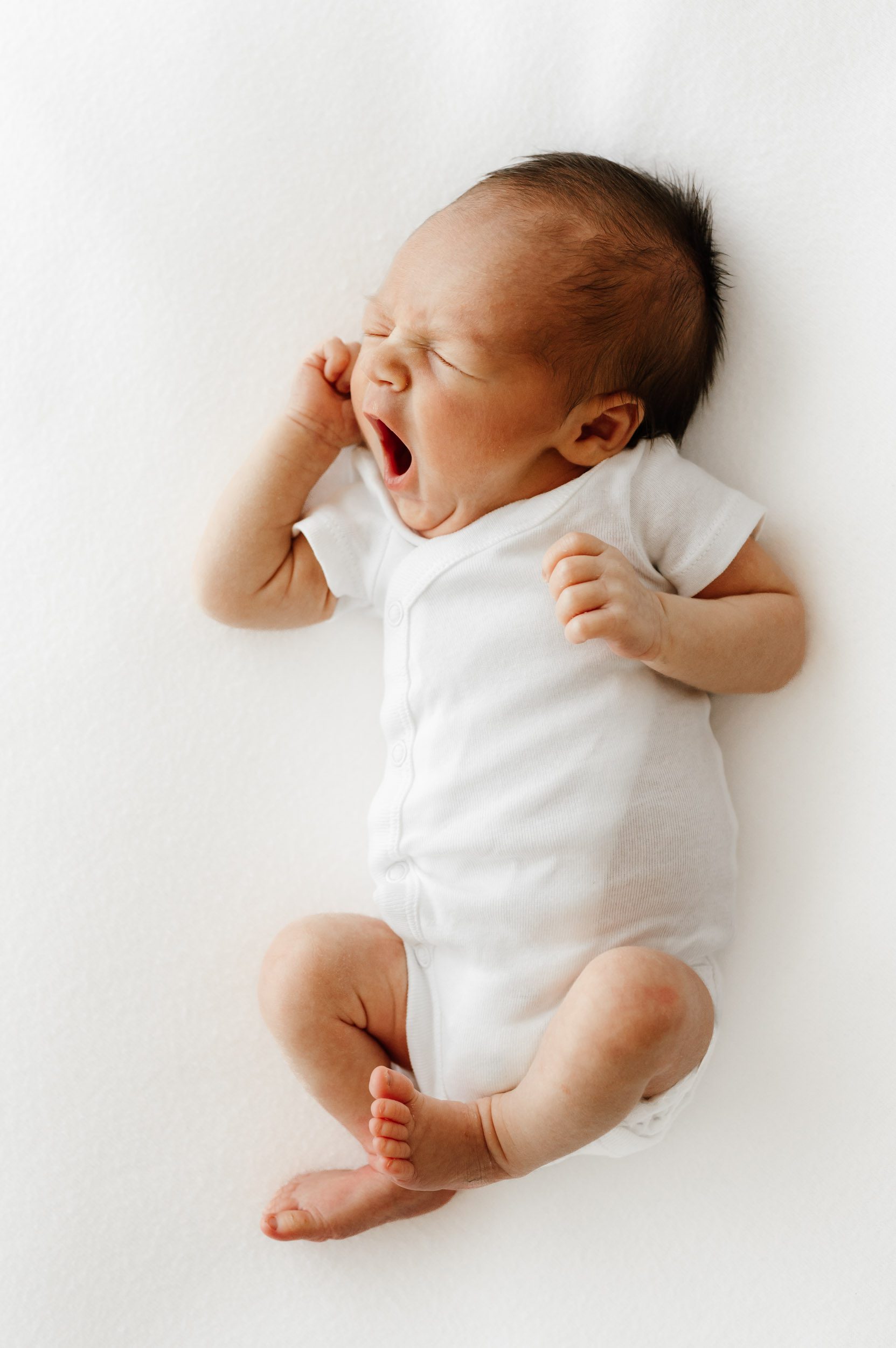 a baby wearing a white onesie laying on a white backdrop holding his hands up to his face and yawning during a newborn photography session