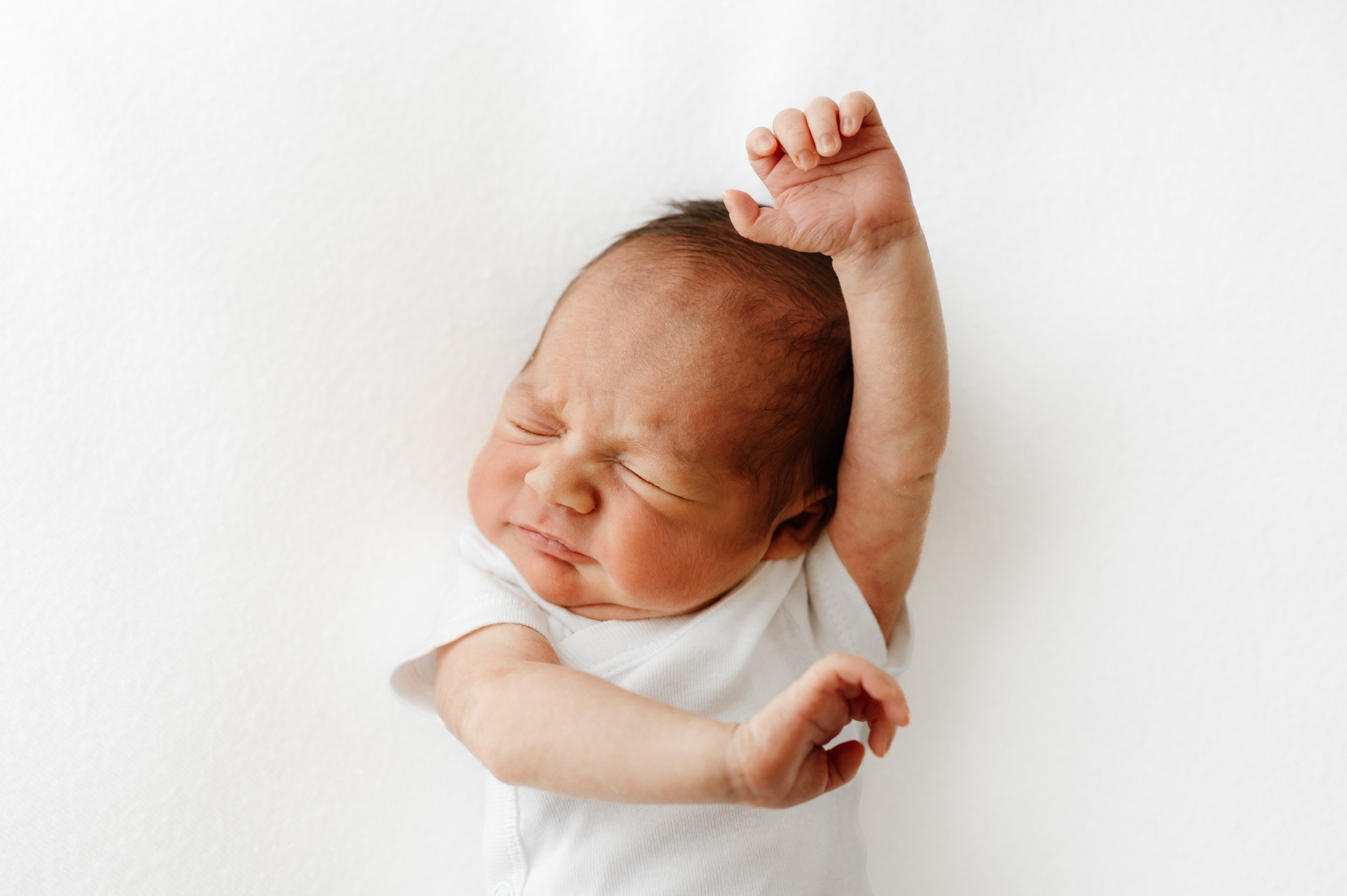 a baby boy wearing a white onesie stretching his arms with his eyes closed during a Pottstown newborn photographer session