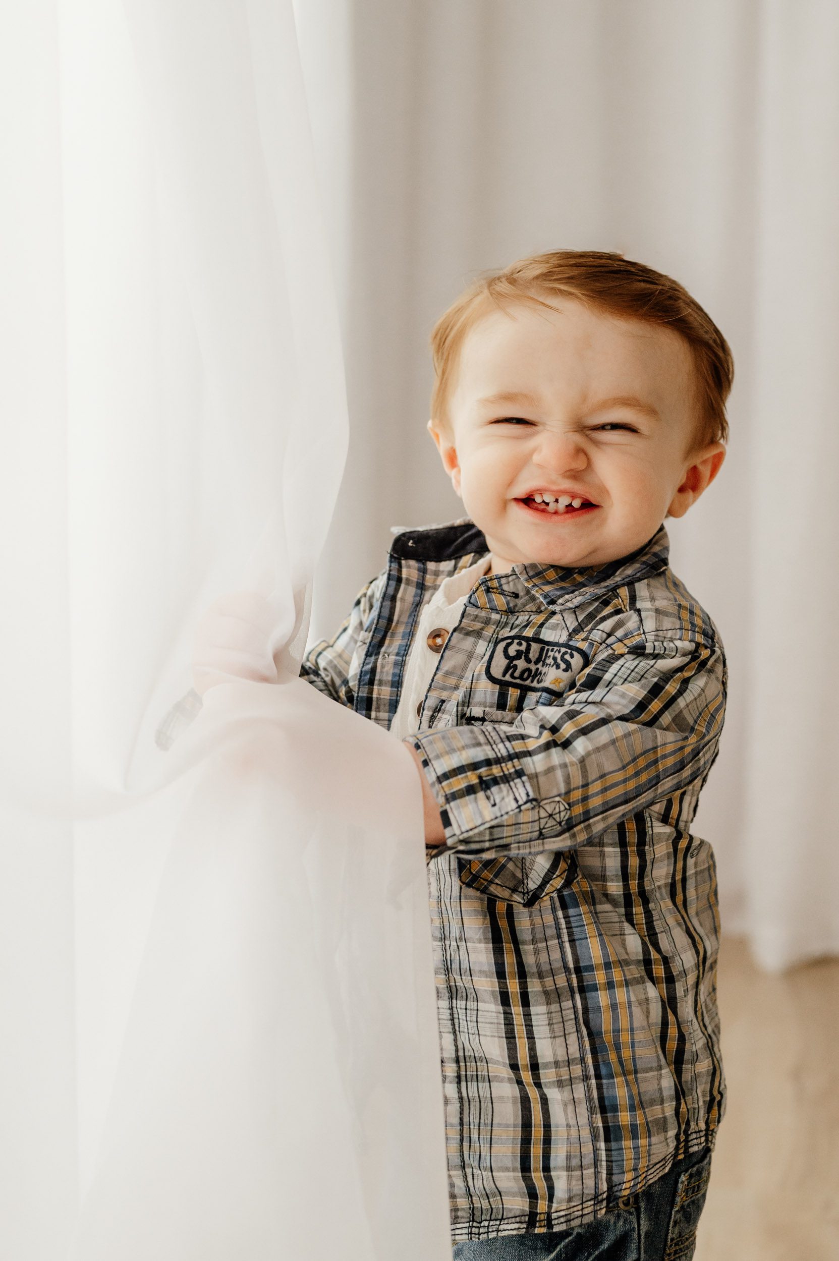 a young boy peeking out from behind a curtain with a huge toothy smile on his face during a 1st birthday photoshoot