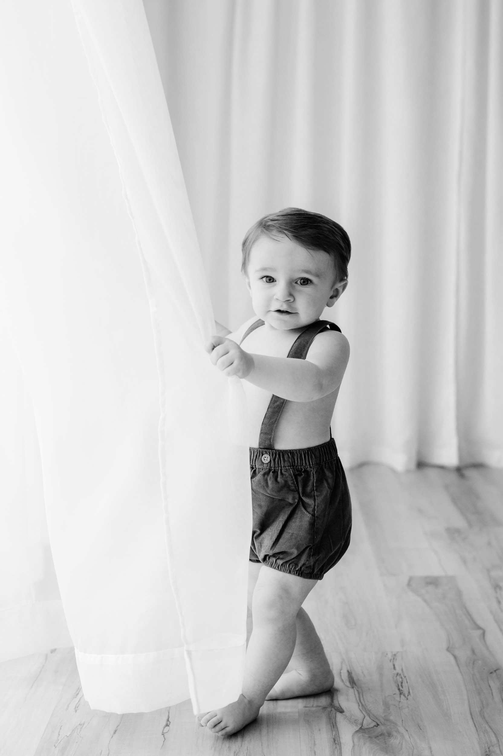 a black and white picture of a young boy looking directly at the camera as he plays with a sheer curtain during a 1st birthday photo session