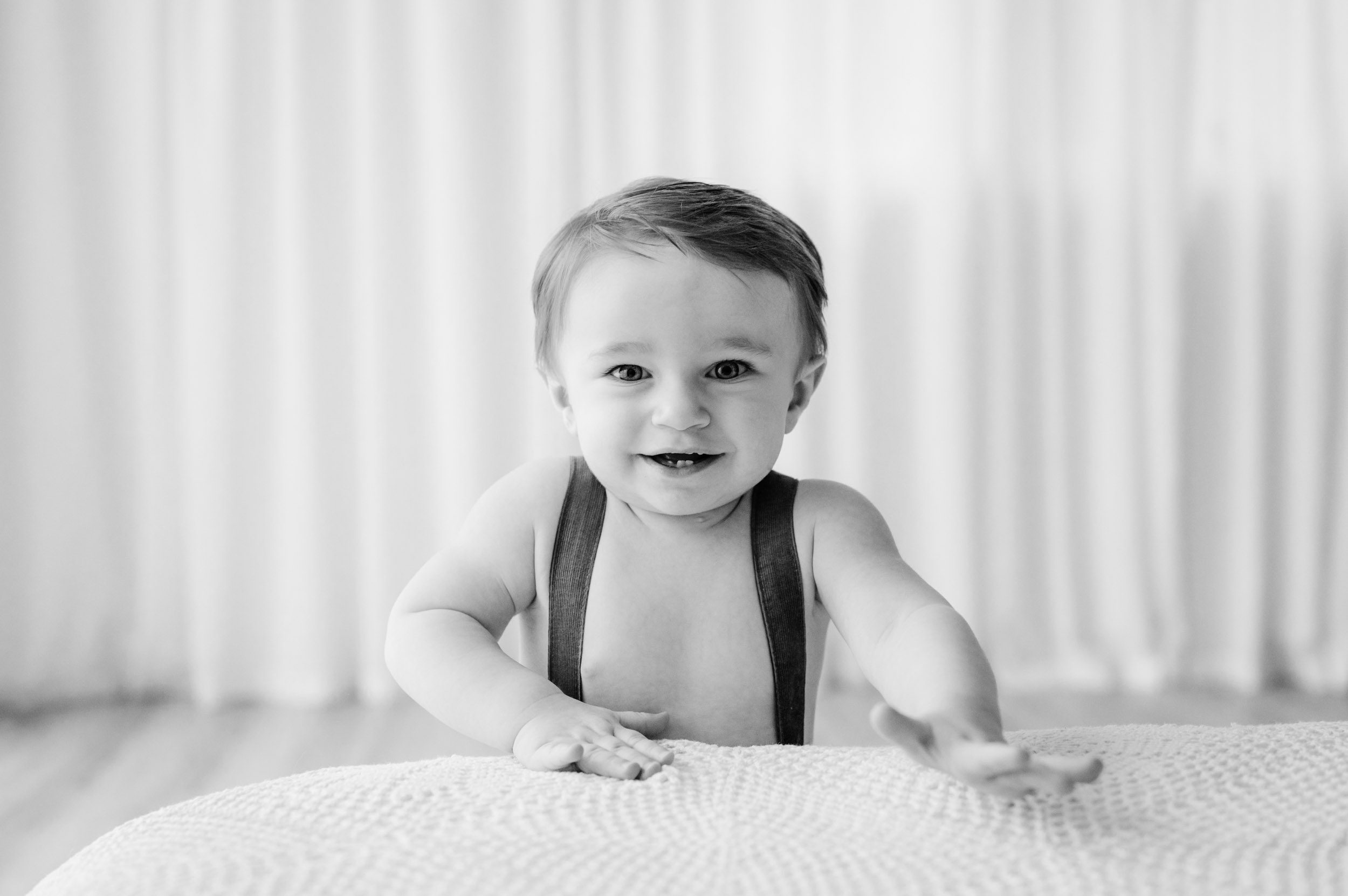 a black and white picture of a young boy standing at the foot of a bed and smiling directly at the camera during a 1st birthday photos session