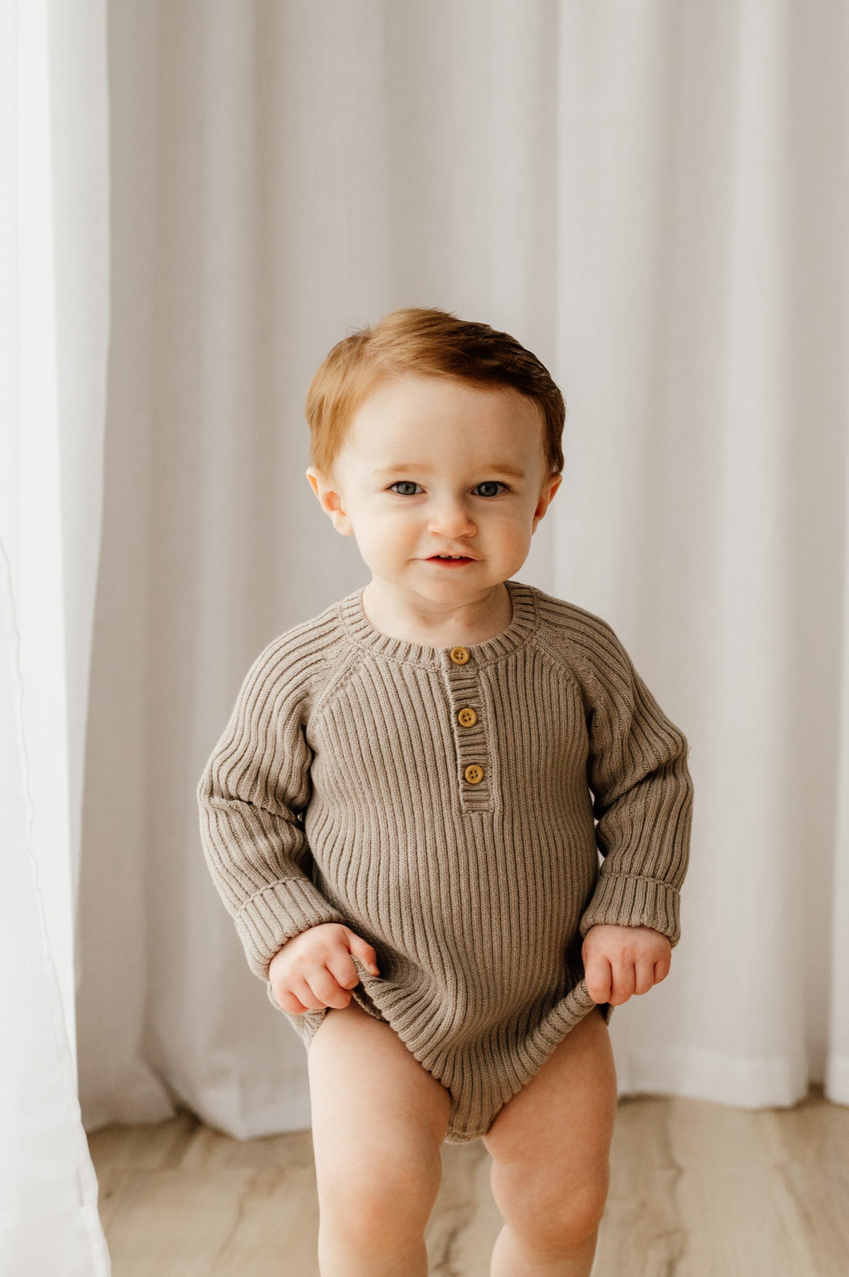 a young boy wearing a beige knit romper walking toward the camera and smiling during a 1st birthday photoshoot