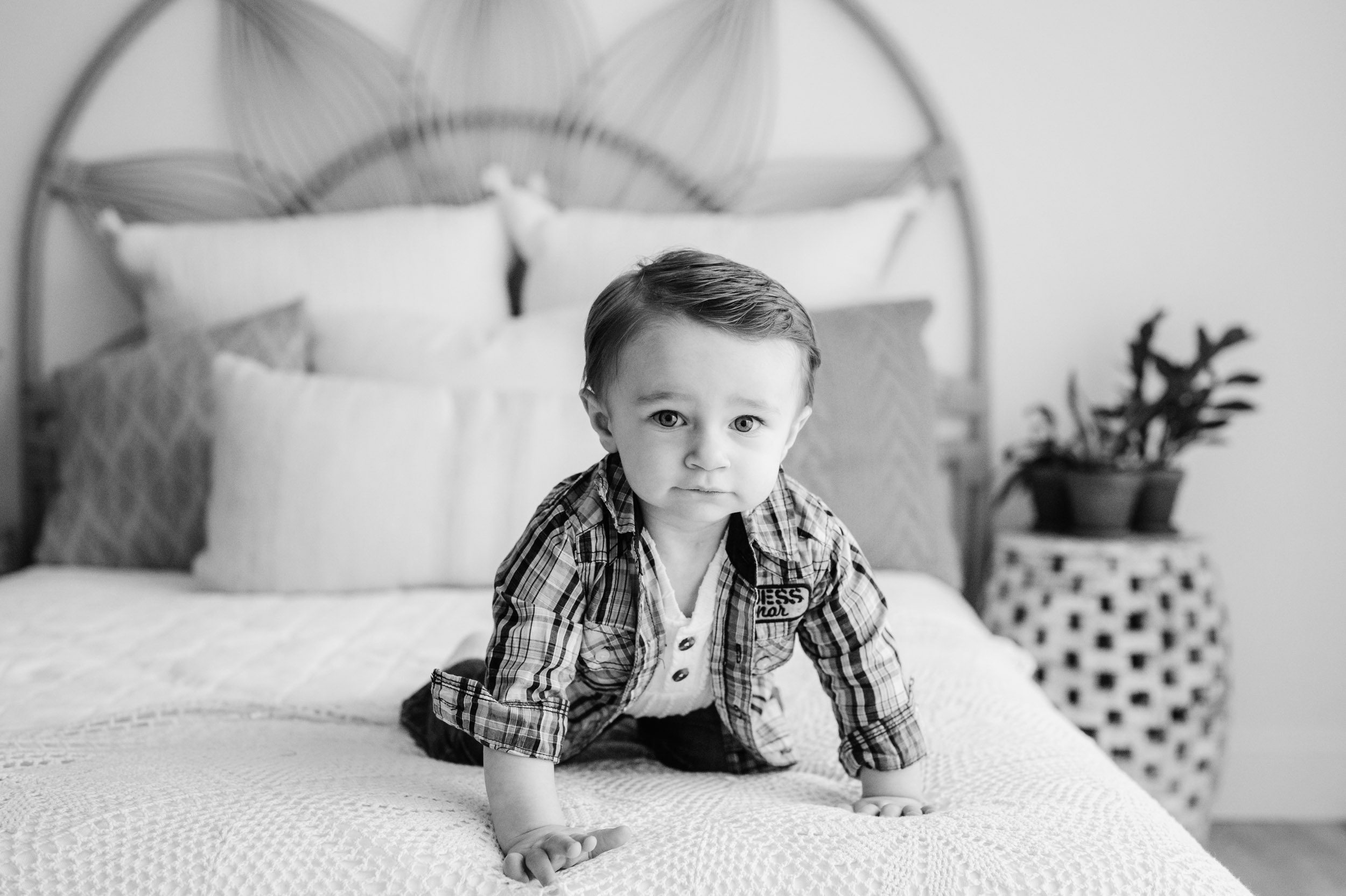 a black and white picture of a young boy crawling across a bed and looking directly at the camera during a 1st birthday photoshoot