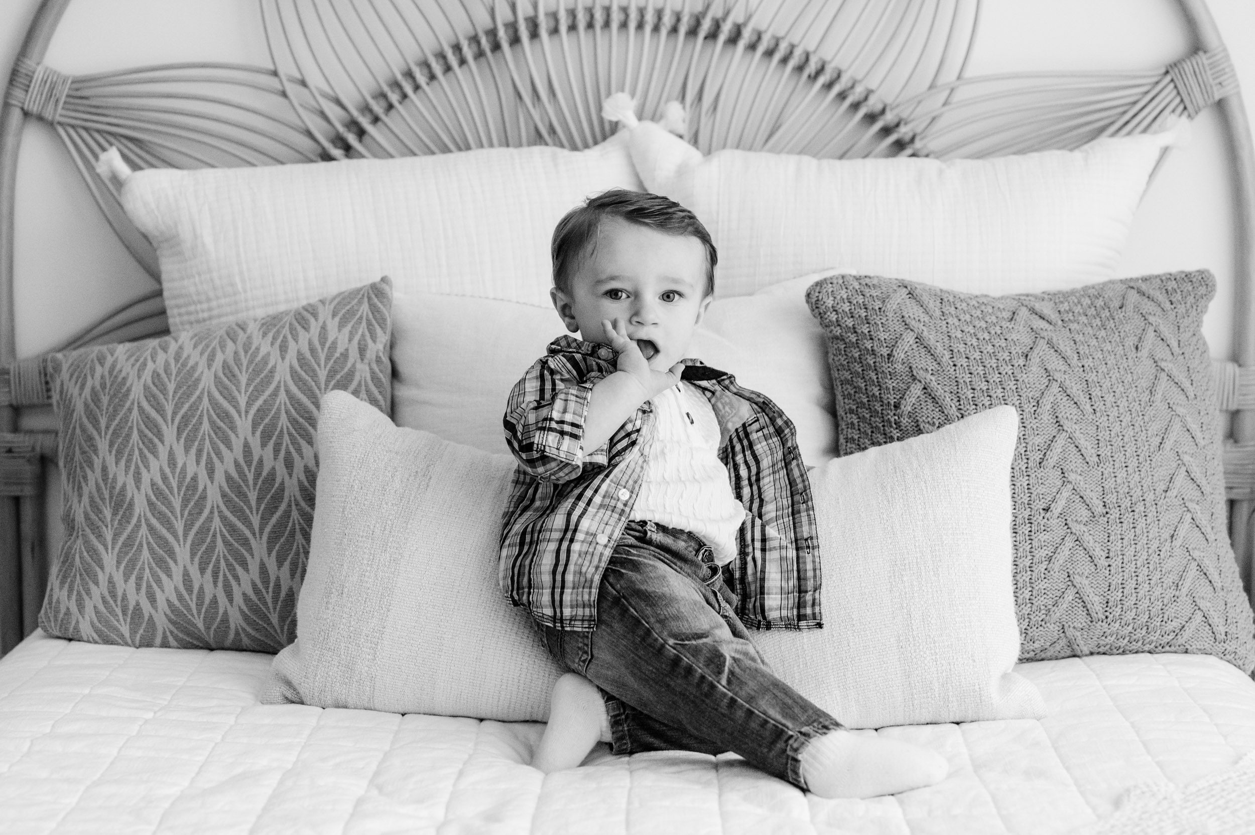 a black and white picture of a young boy sitting on a bed and leaning against the pillow as he sucks on his finger and looks right at the camera 