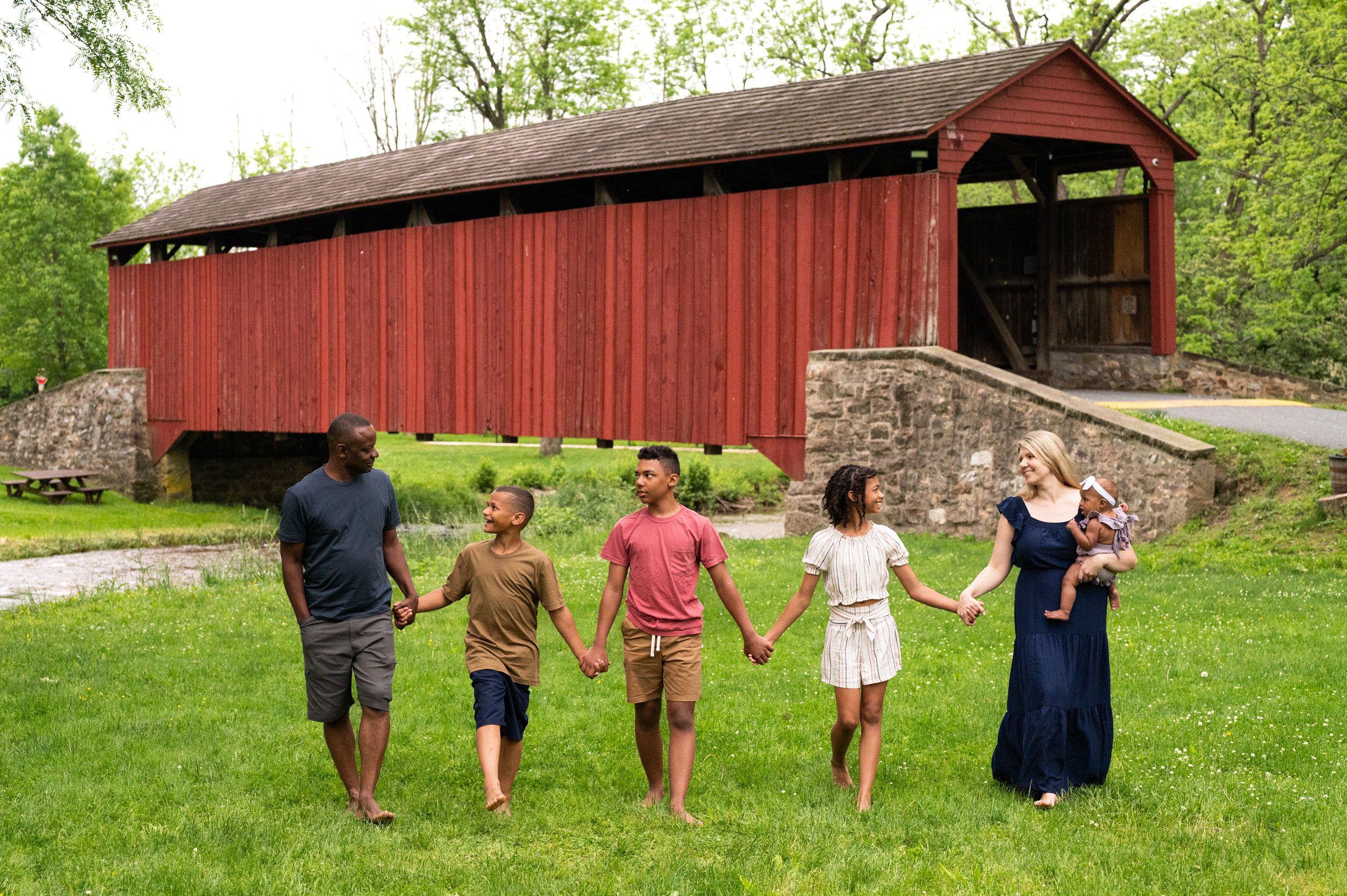 a family of six walking through the grass holding hands and smiling at the other with a big red covered bridge in the background during a fall family photoshoot