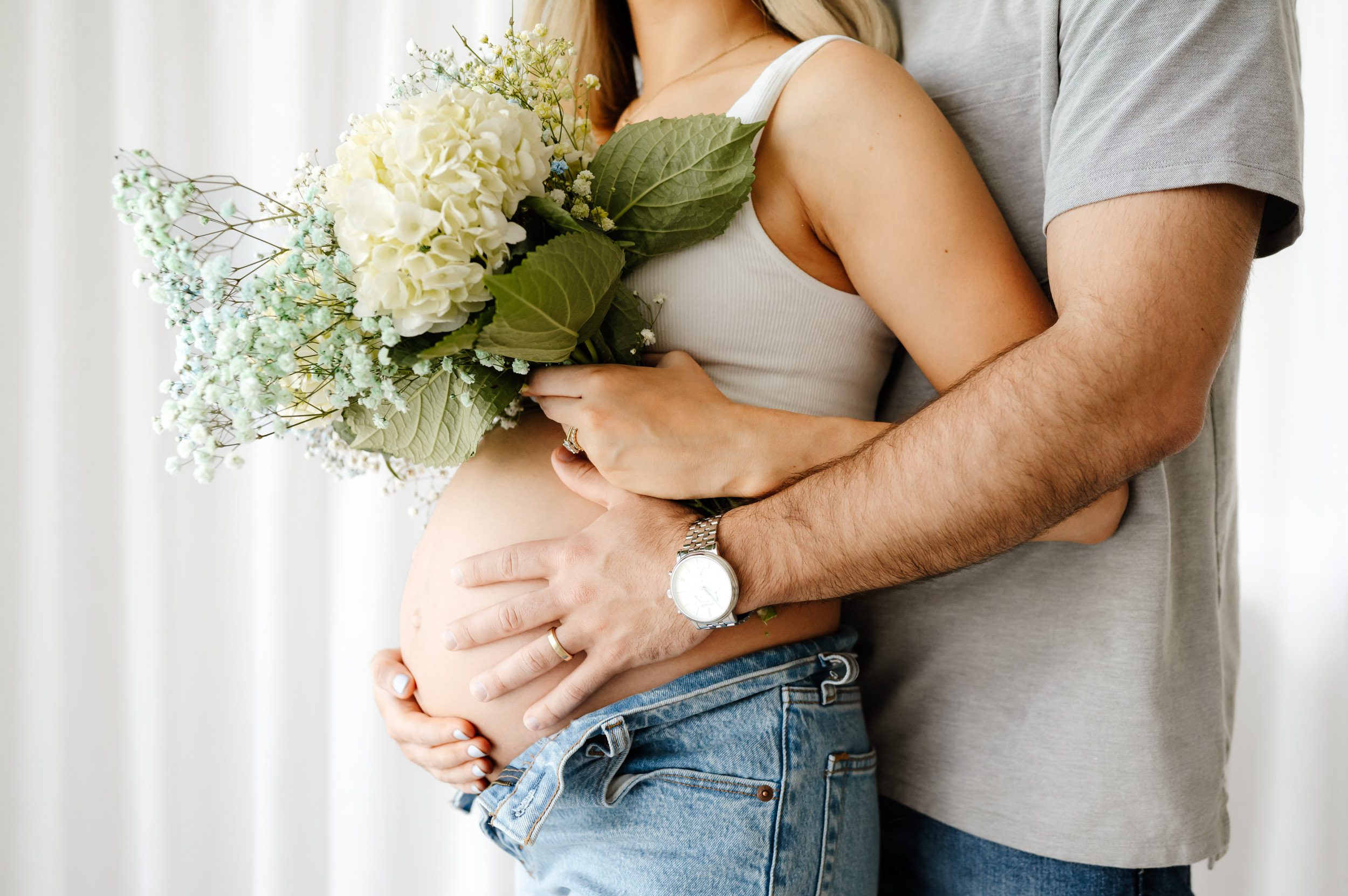 a close up picture of an expecting mom's bare belly as she holds a bouquet of flowers in her hand and dad hugs her from behind during a lifestyle maternity photoshoot