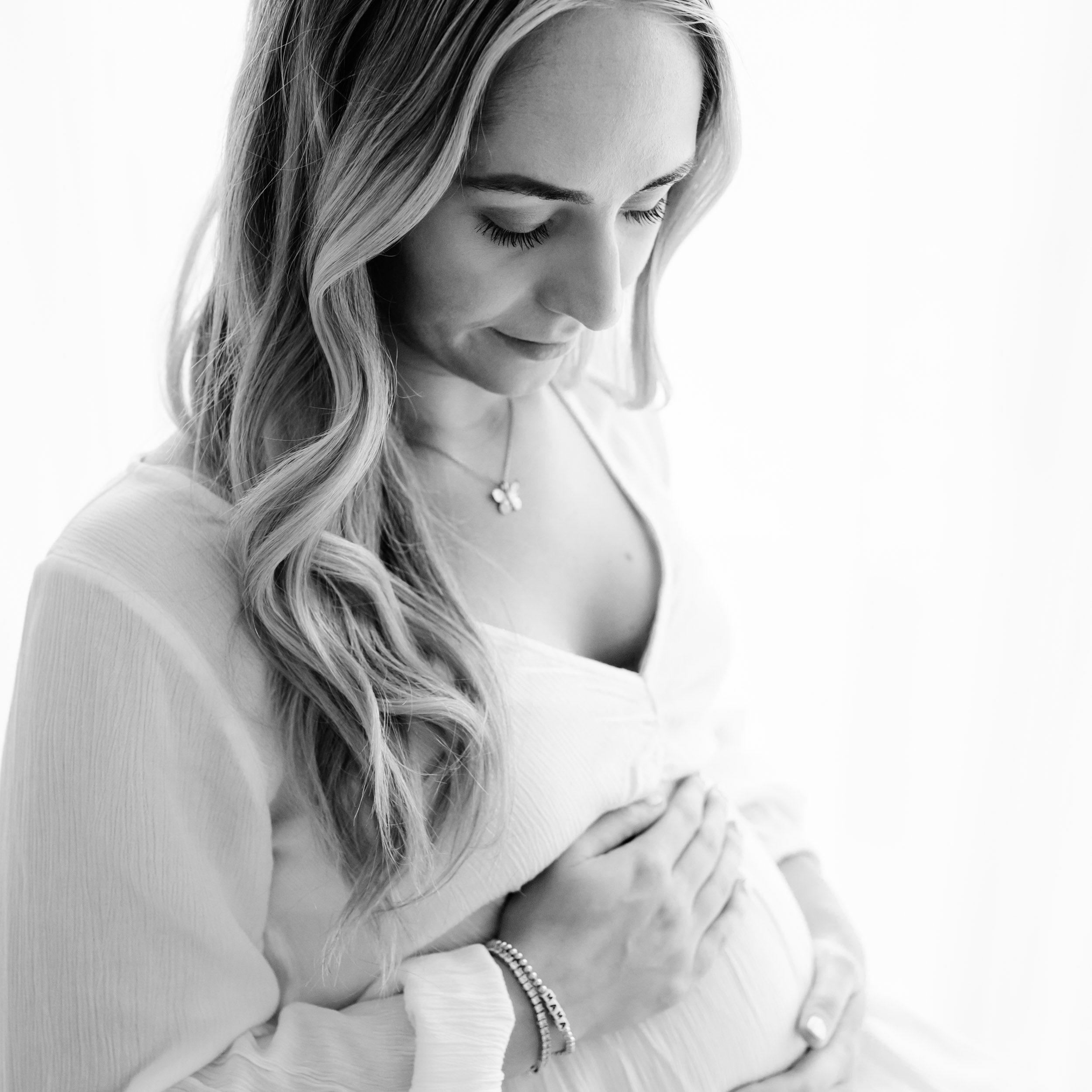 a black and white backlit photo of an expecting mom wearing a white dress and smiling down as she cradles her belly with her hands