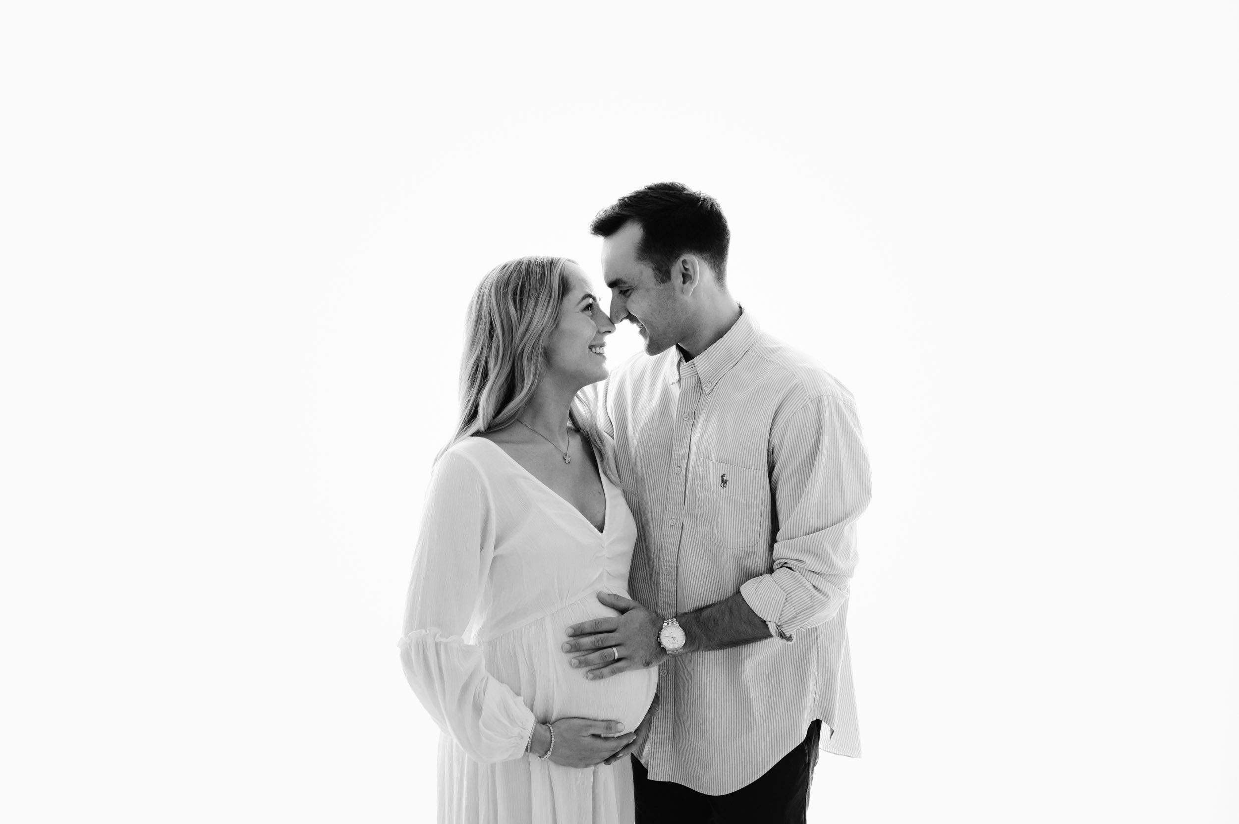 a black and white backlit photo of expecting parents standing in front of a huge window with their hands on mom's belly as they smile at each other and touch their noses together
