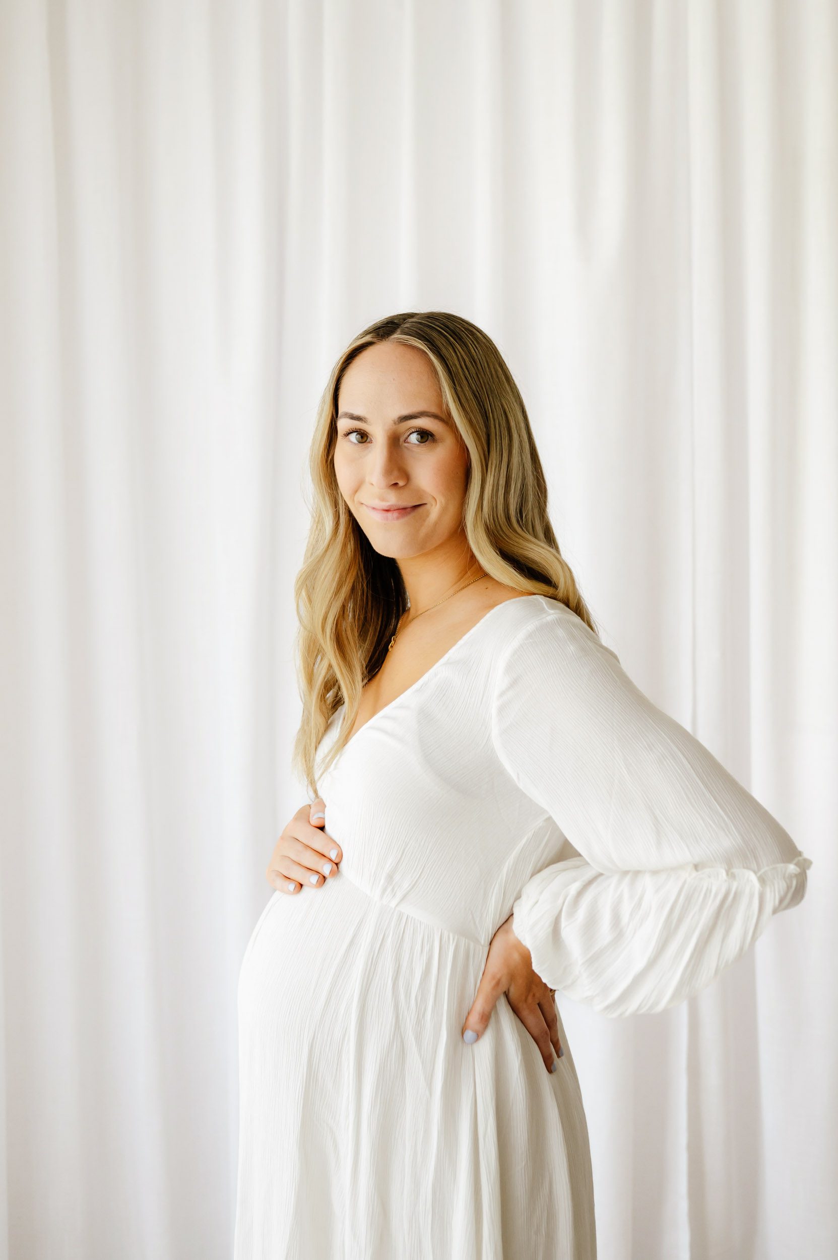 an expecting mom wearing a white dress touching her belly and smiling at the camera during a maternity photos session
