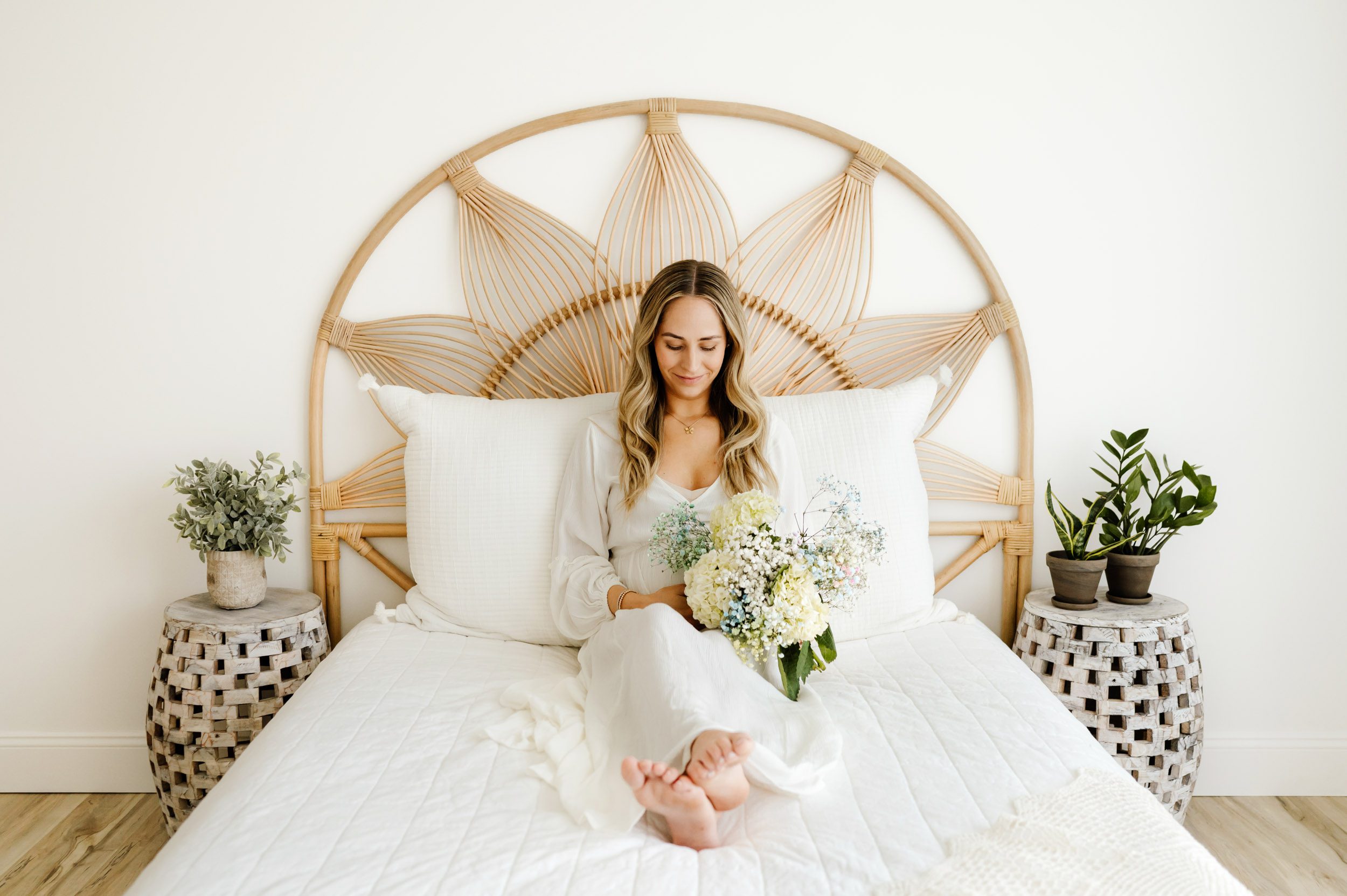 an expecting mom wearing a white dress sitting on a bed holding a bouquet of flowers and smiling down at her belly during a maternity photos session