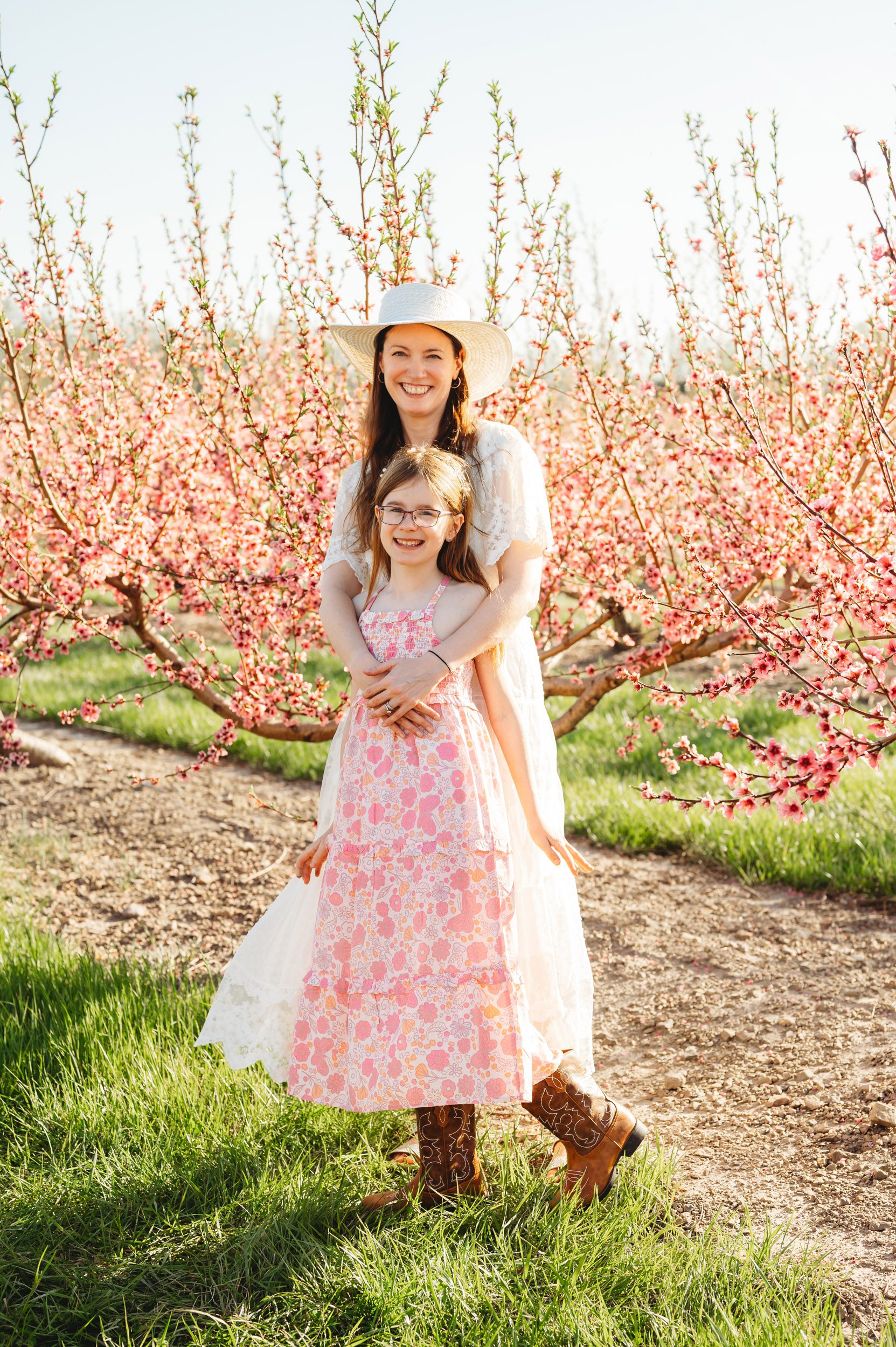 a mom hugging her daughter as they both smile at the camera during a mini session in an orchard full of pink peach blossoms in bloom