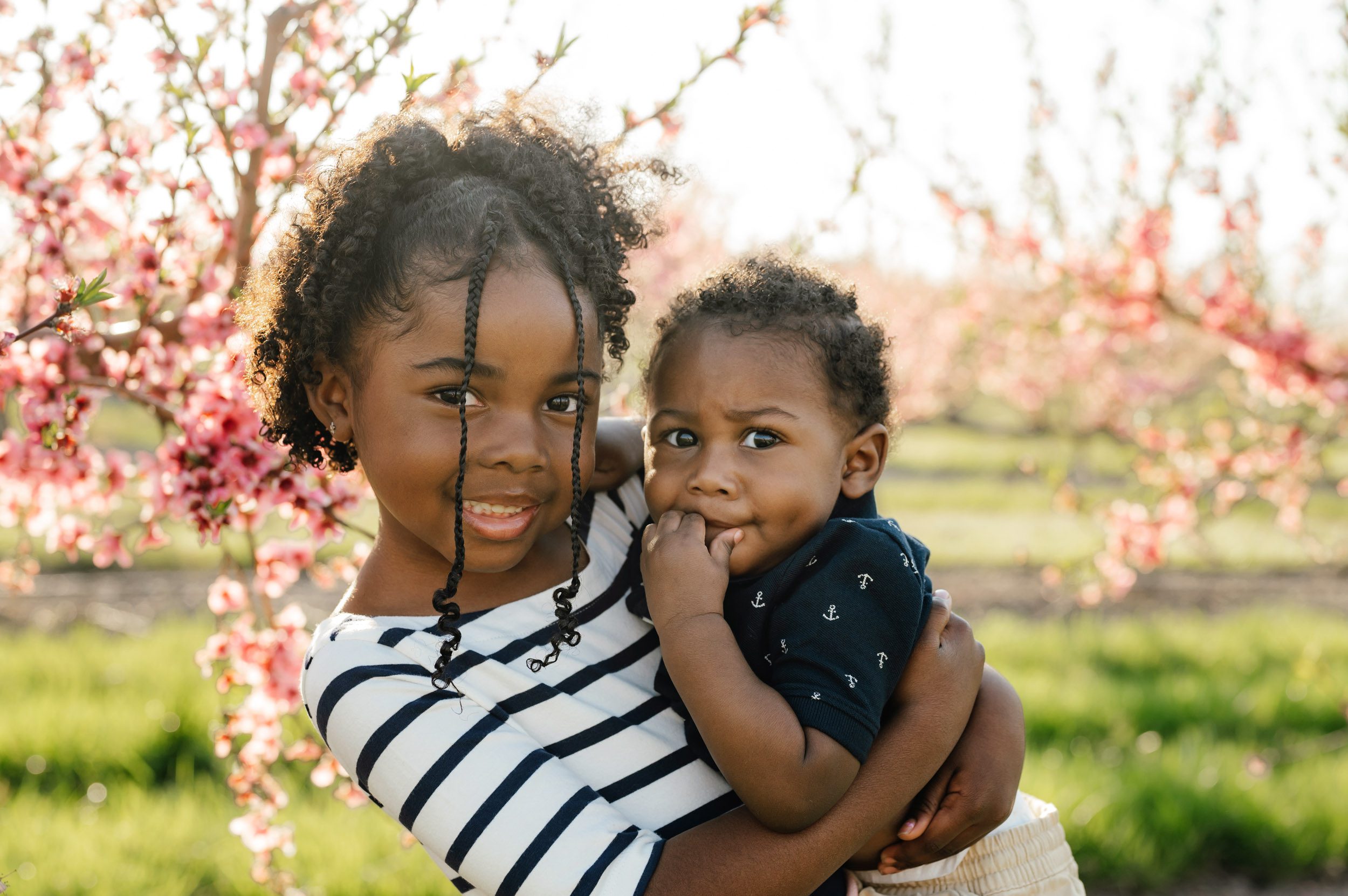 a girl holding her little brother in her arms as they both look at the camera and smile during a mini session in an orchard full of pink peach blossoms in bloom