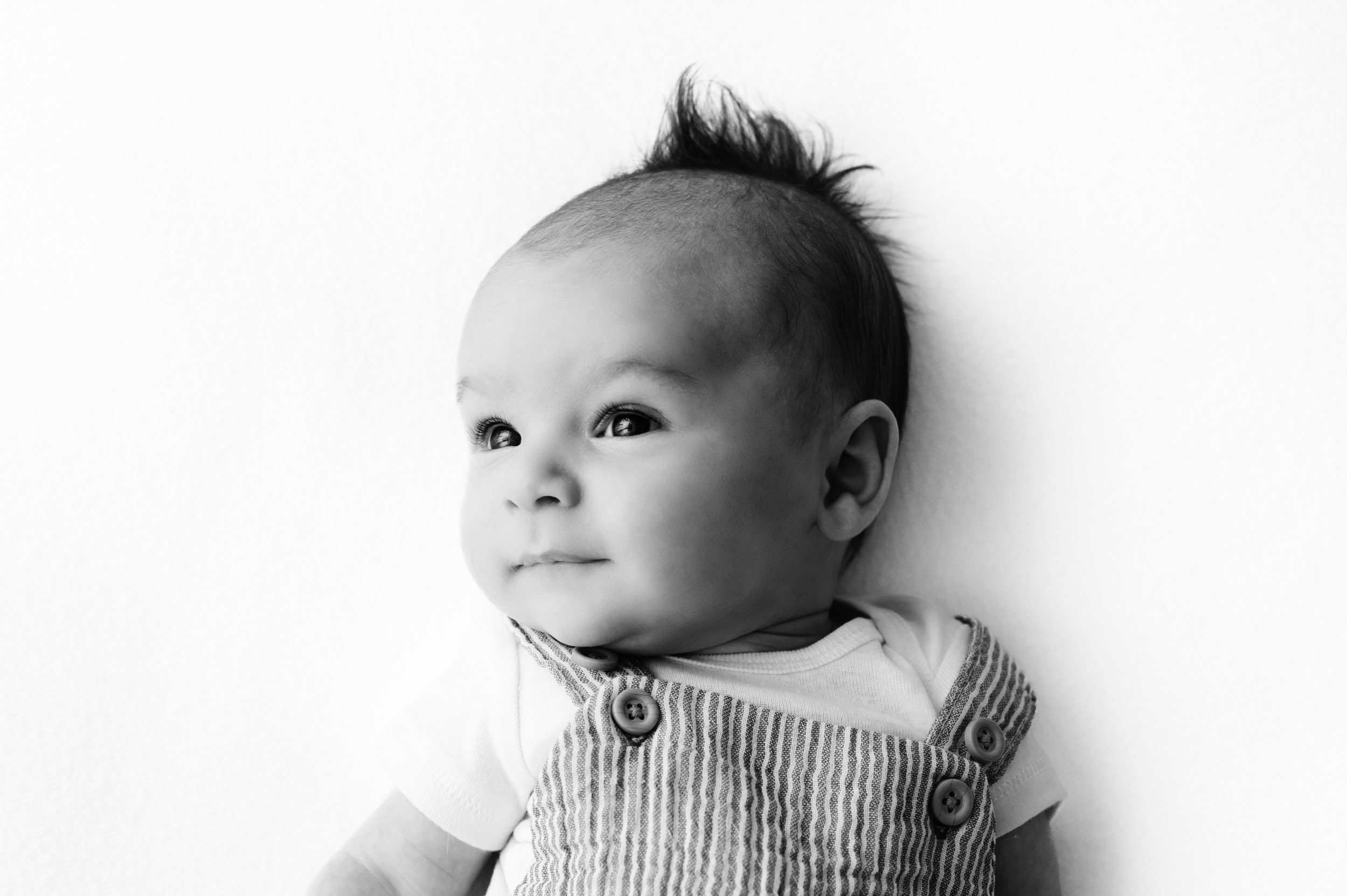 a black and white picture of a baby boy wearing a striped onesie laying on a white backdrop as he looks off toward the window to his side during a newborn photoshoot