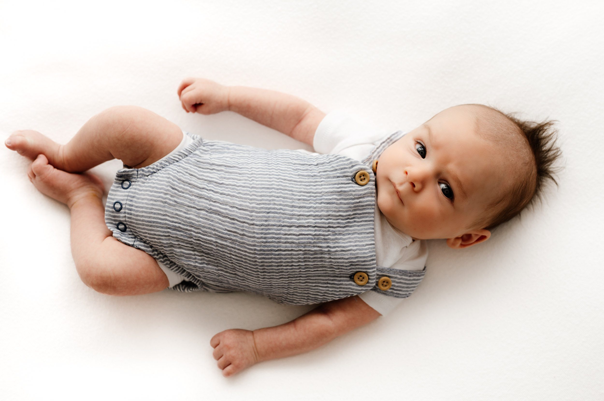 a baby boy wearing a blue and white striped romper laying on a white bean bag and staring intently up at the camera with his head tilted to one side during a newborn photographer session