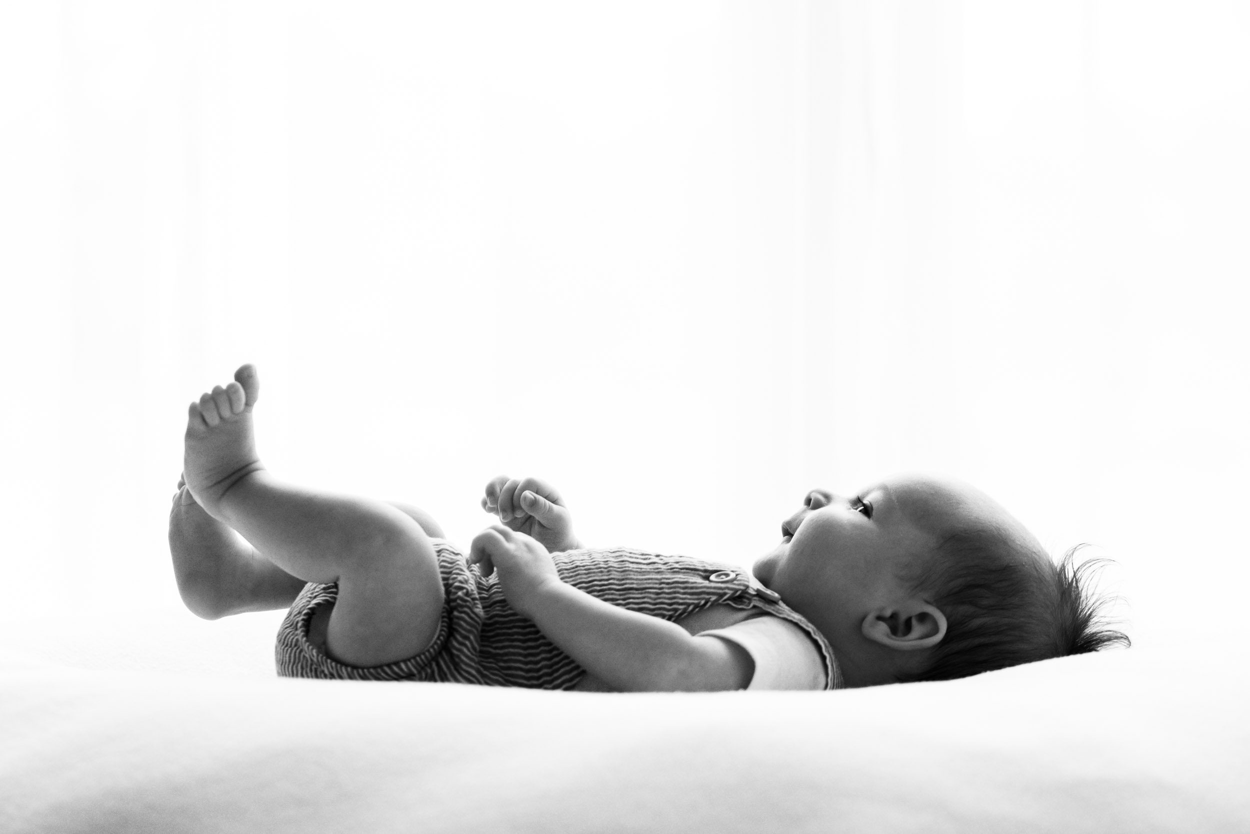 a backlit black and white picture of a baby boy laying on a white bean bag and lifting his feet up in the air during a Pottstown newborn photographer session