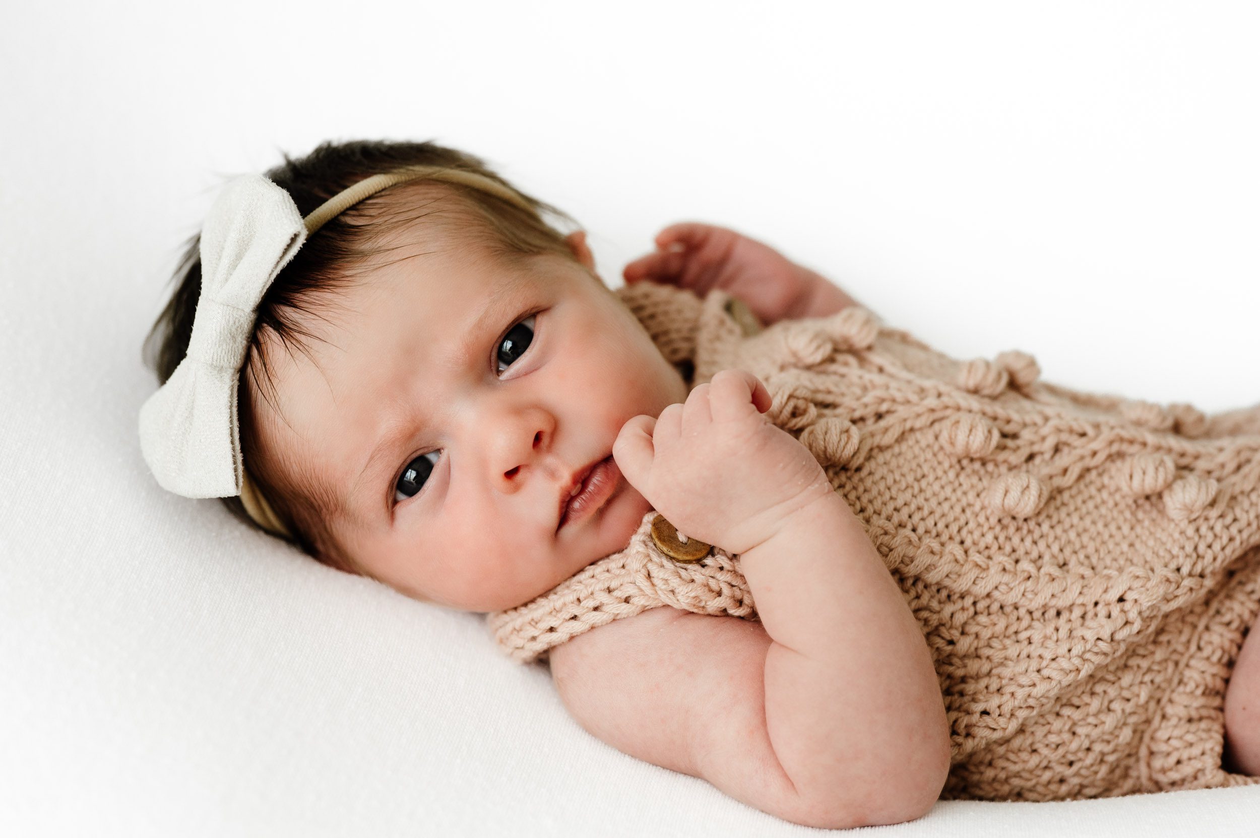 a baby girl waring a light pink knit romper and white headband laying on a white backdrop and touching her finger to her cheek as she looks directly at the camera during a natural newborn studio photoshoot