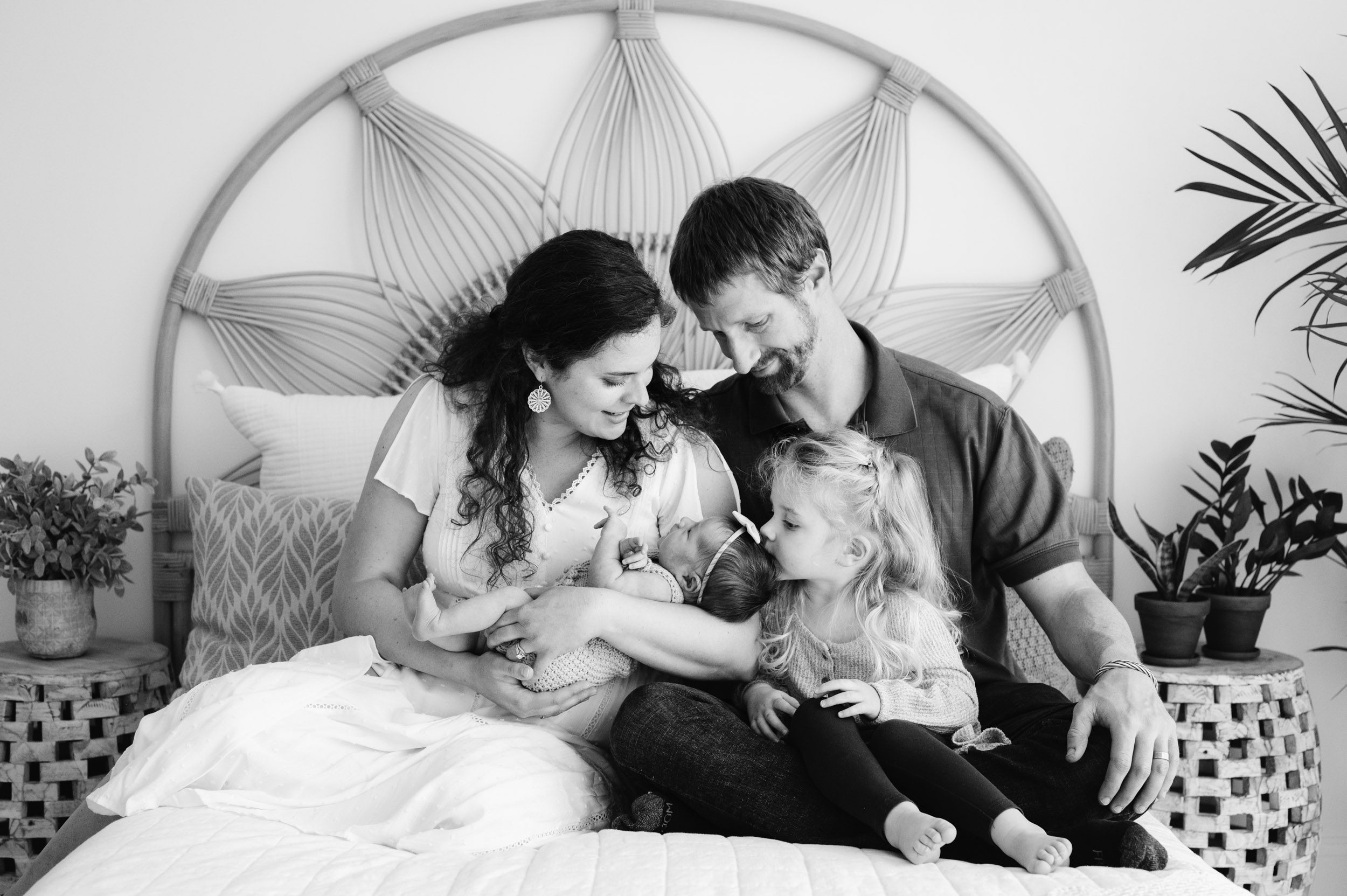 a black and white picture of a family of four sitting on a bed as mom cradles her newborn daughter in her arms while big sister sits on dad's lap and kisses the baby on the head during a studio newborn photo session