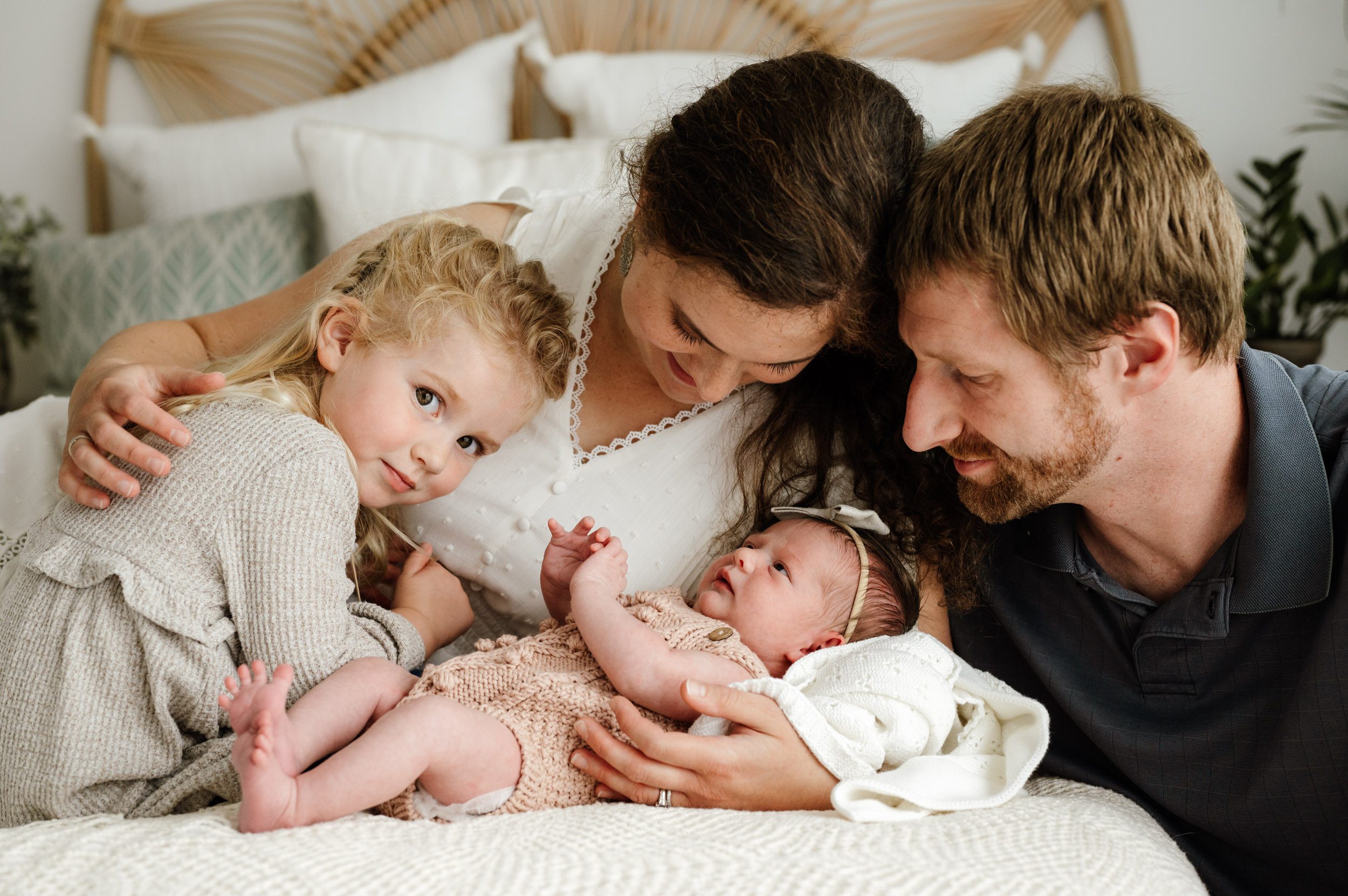 A new family of four laying on the bed with their baby girl laying on the bed in front of mom and big sister looking directly at the camera with a hint of a smile on her face during a natural newborn photo session