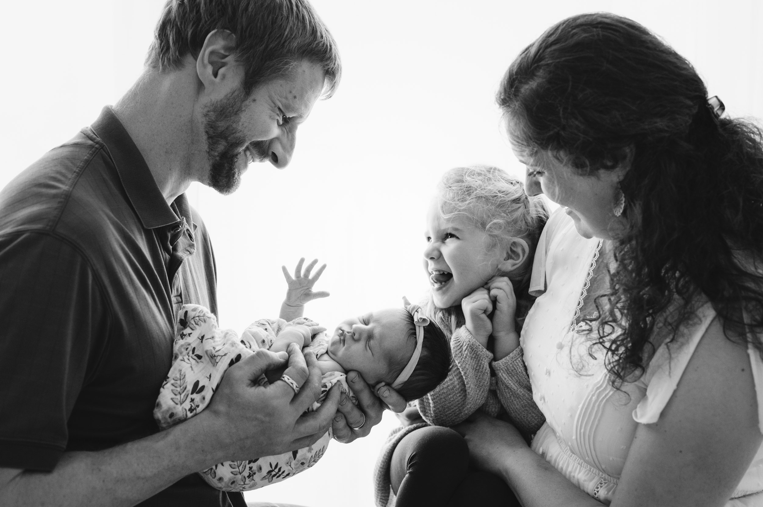 a black and white back lit picture of a dad holding his newborn daughter in his arms and smiling down at her while mom holds her older daughter who is sticking her tongue at her dad during a studio newborn photoshoot